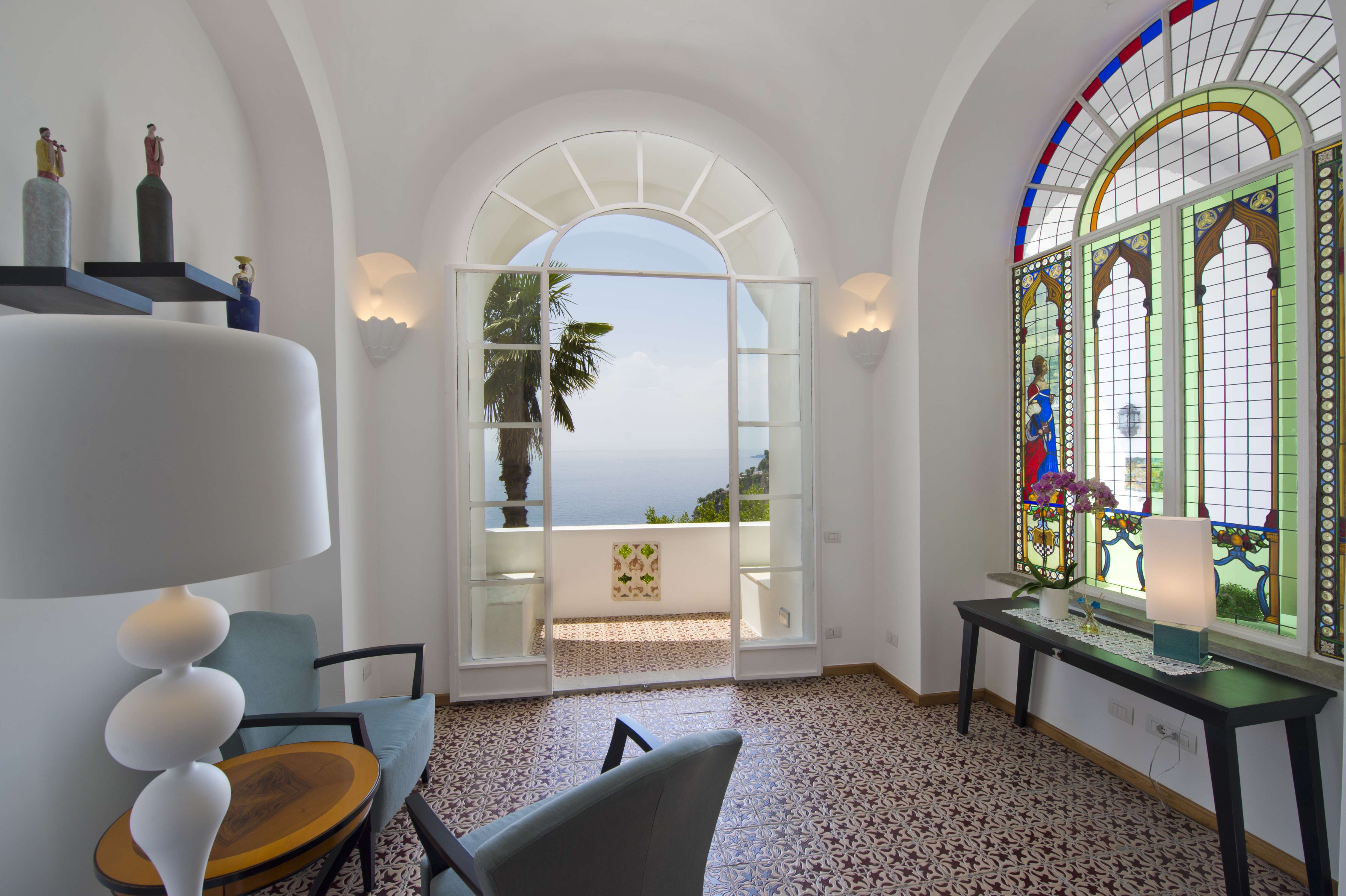 Exclusive Resorts' Amalfi villa interior with arched windows, stained glass, and a balcony overlooking the Mediterranean Sea.