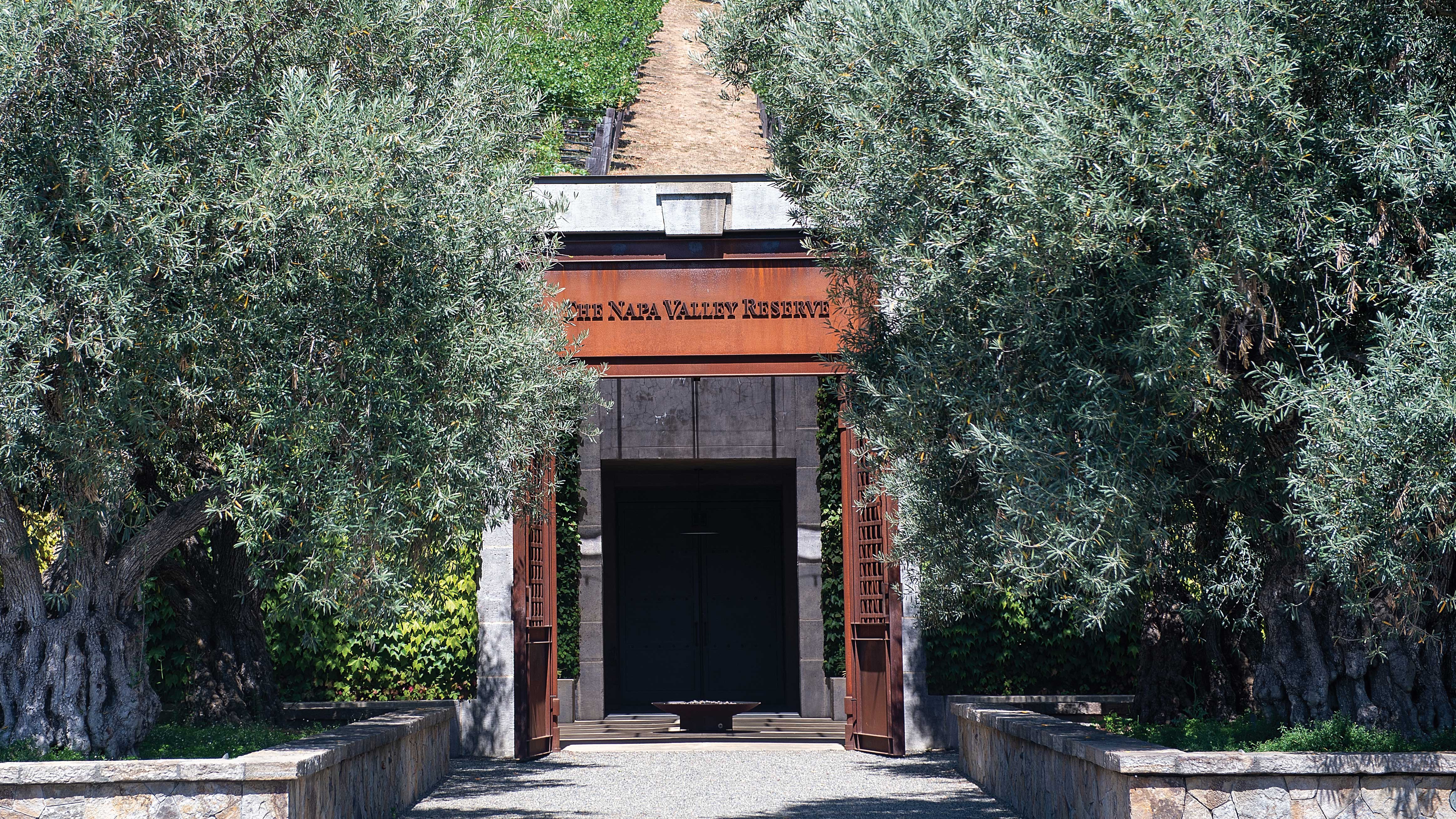 Entrance to The Napa Valley Reserve covered by lush olive trees, with a bold sign over the doorway inviting visitors into the prestigious vineyard.