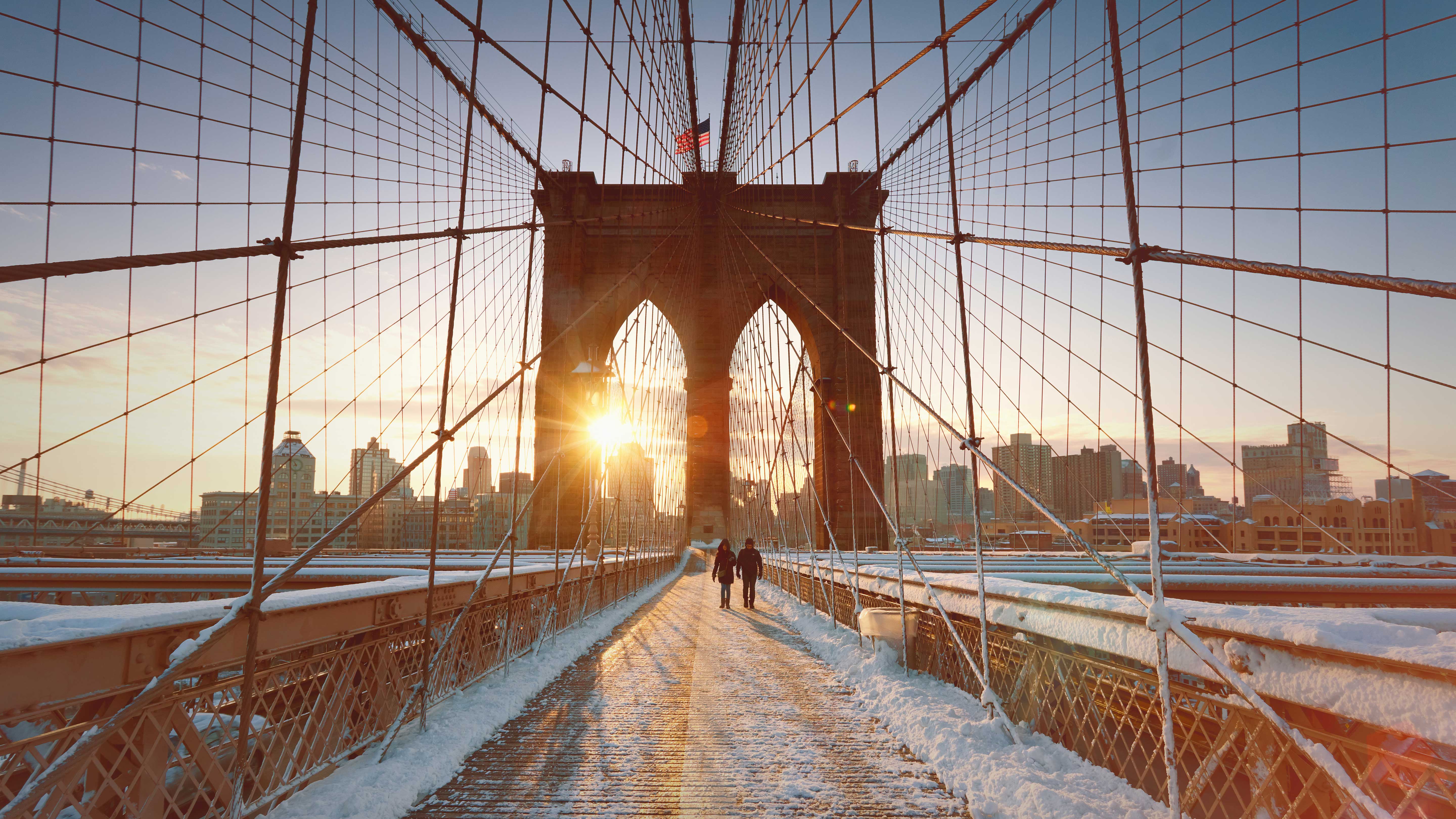 Winter sunrise casting over the snow-covered Brooklyn Bridge with pedestrians strolling along the pathway.