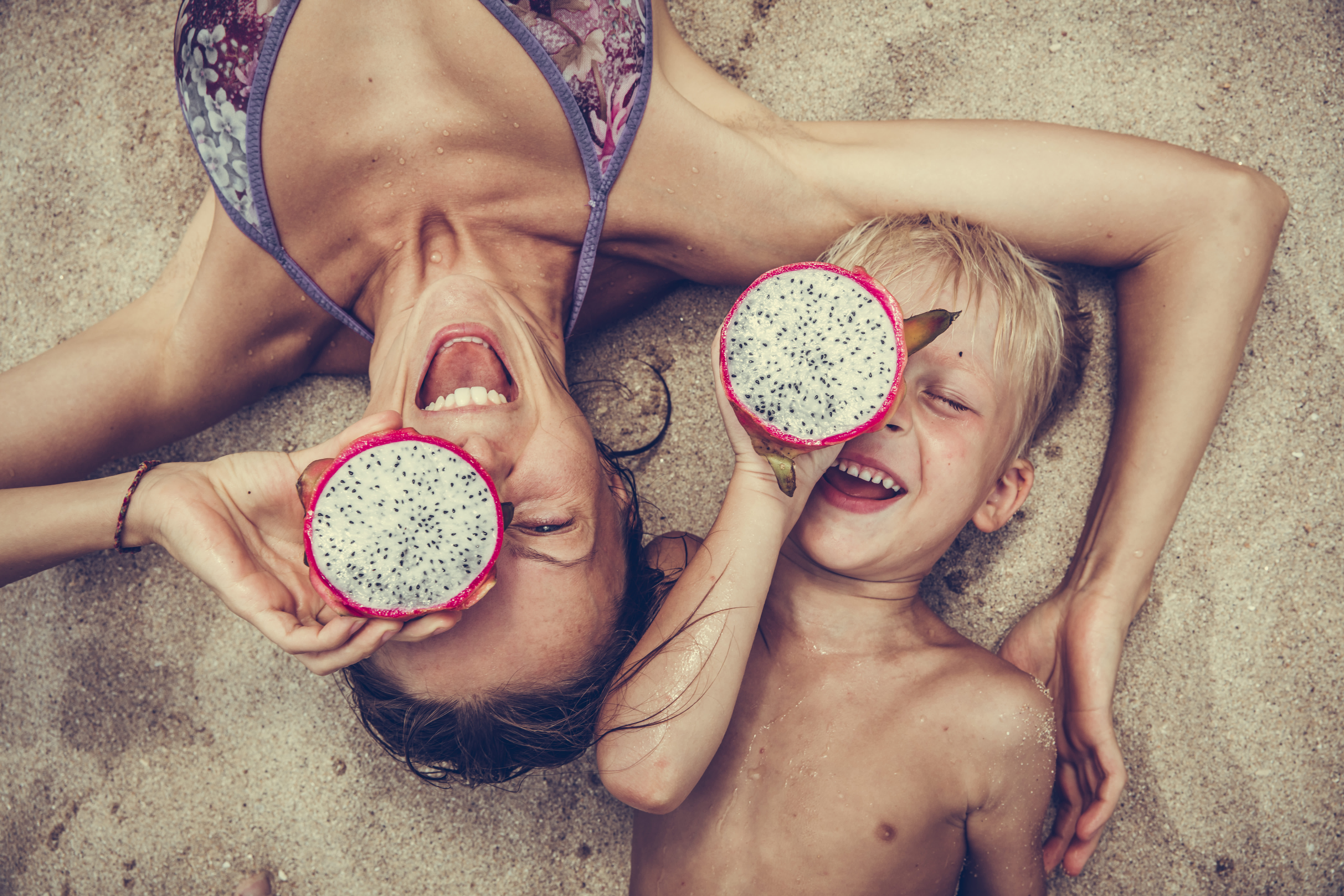 Woman and child laughing on the beach holding dragon fruit slices over their eyes.