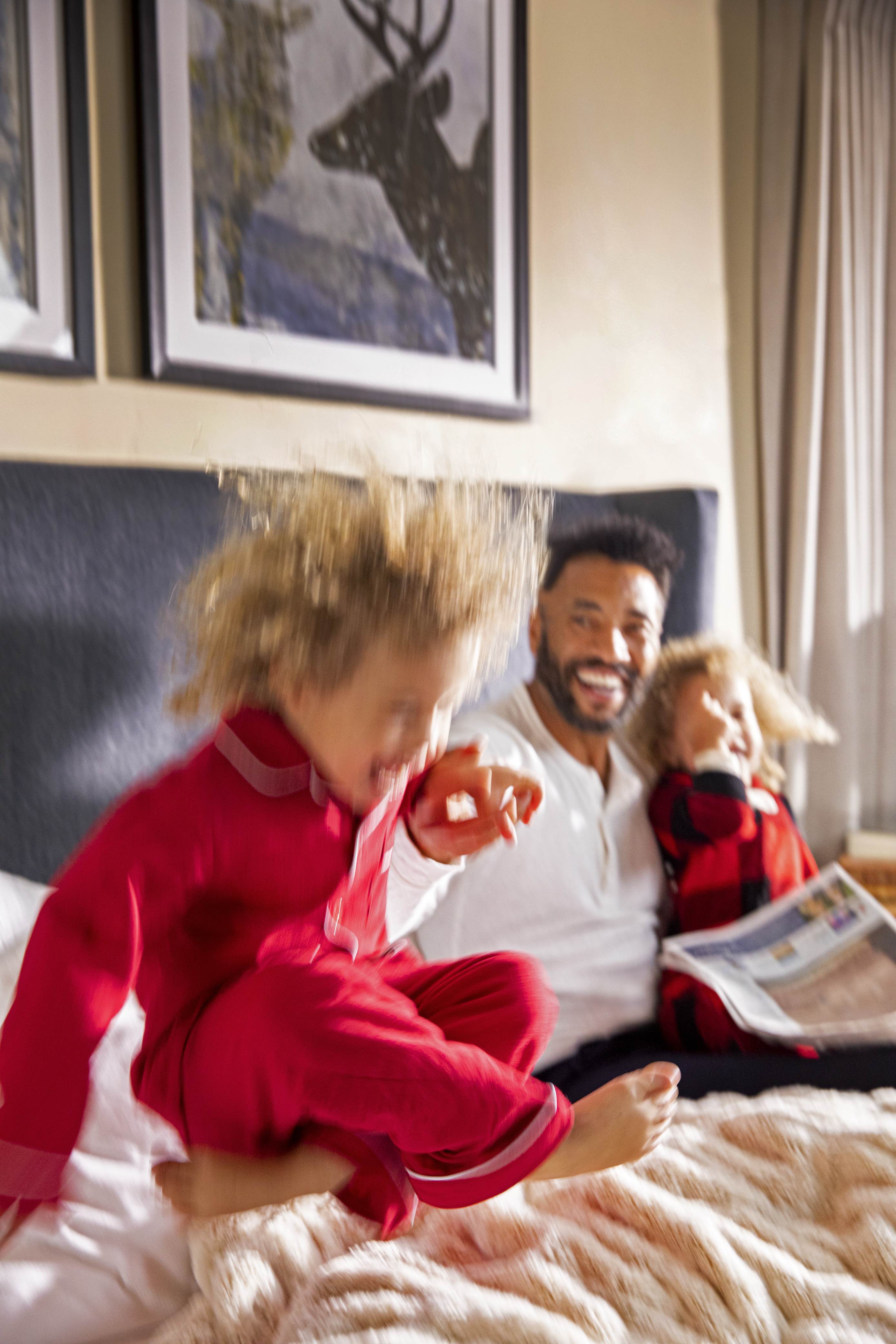 A kid jumping on a bed while the father and brother laugh.