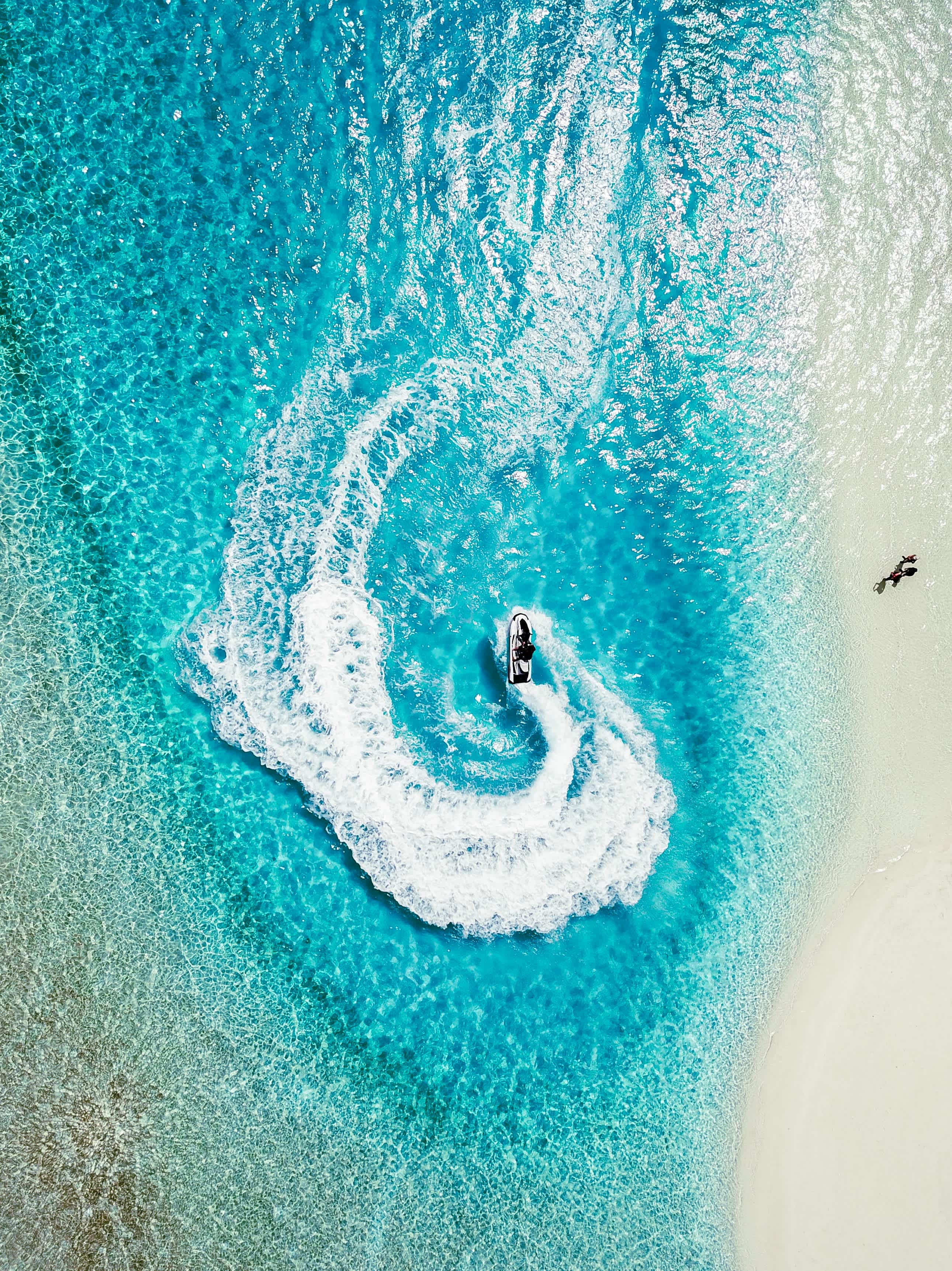 Aerial view of a jet ski making a sharp turn in the clear turquoise waters near a sandy beach in the Maldives.