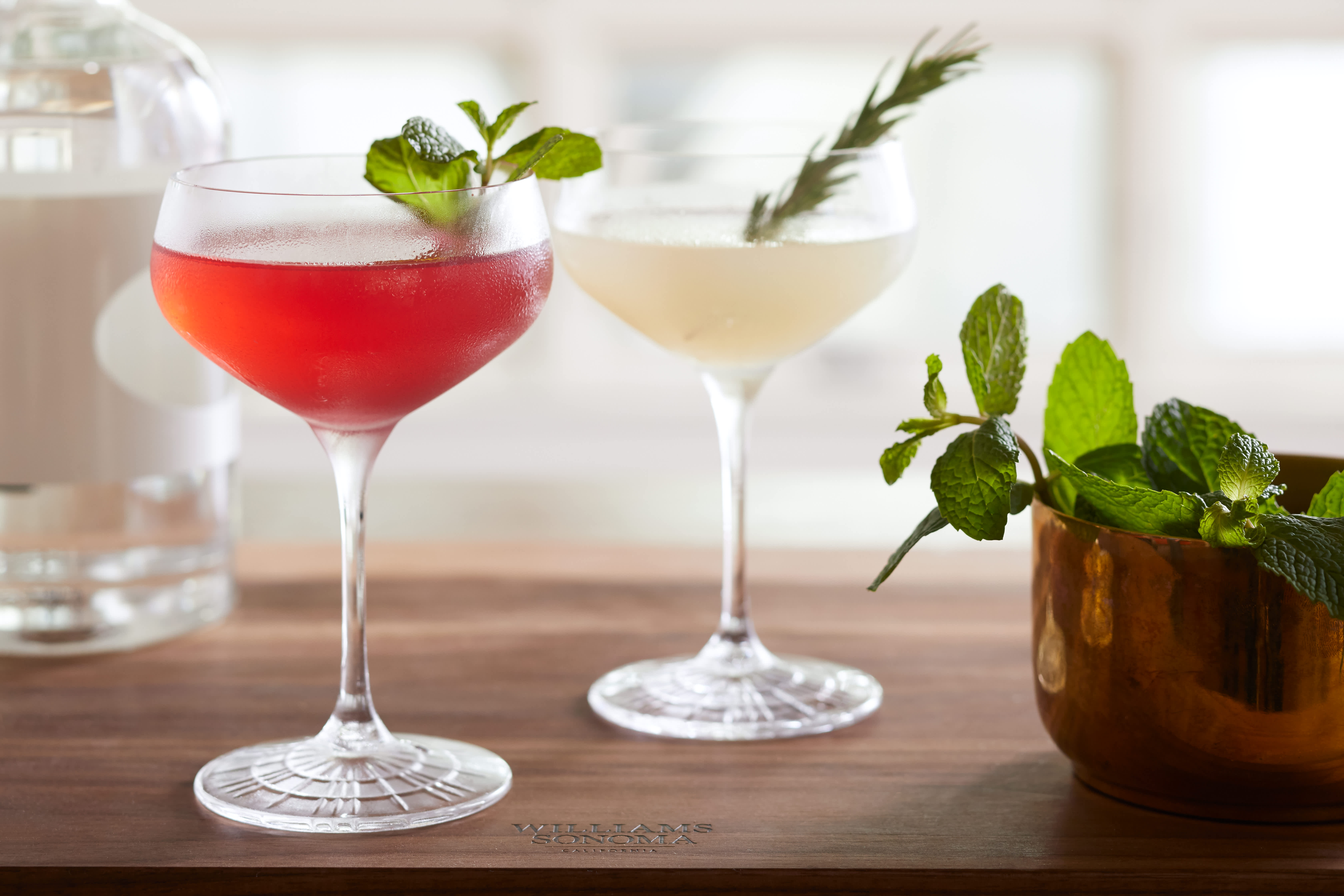 Two elegant stemmed cocktail glasses, one with a red beverage garnished with mint, another with a clear beverage and rosemary.