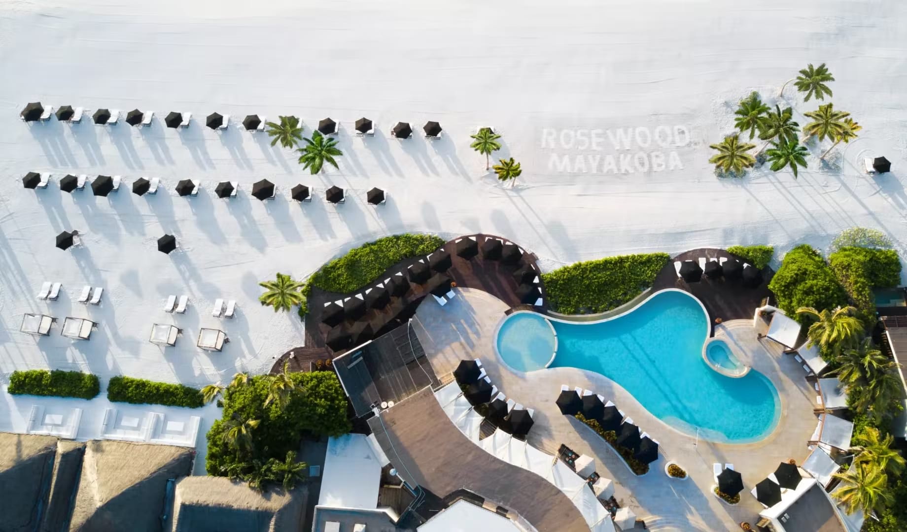 Aerial view of the Rosewood Mayakoba's distinctive pool alongside a perfectly arranged row of beach cabanas and palm trees, close to Exclusive Resorts properties in Riviera Maya.