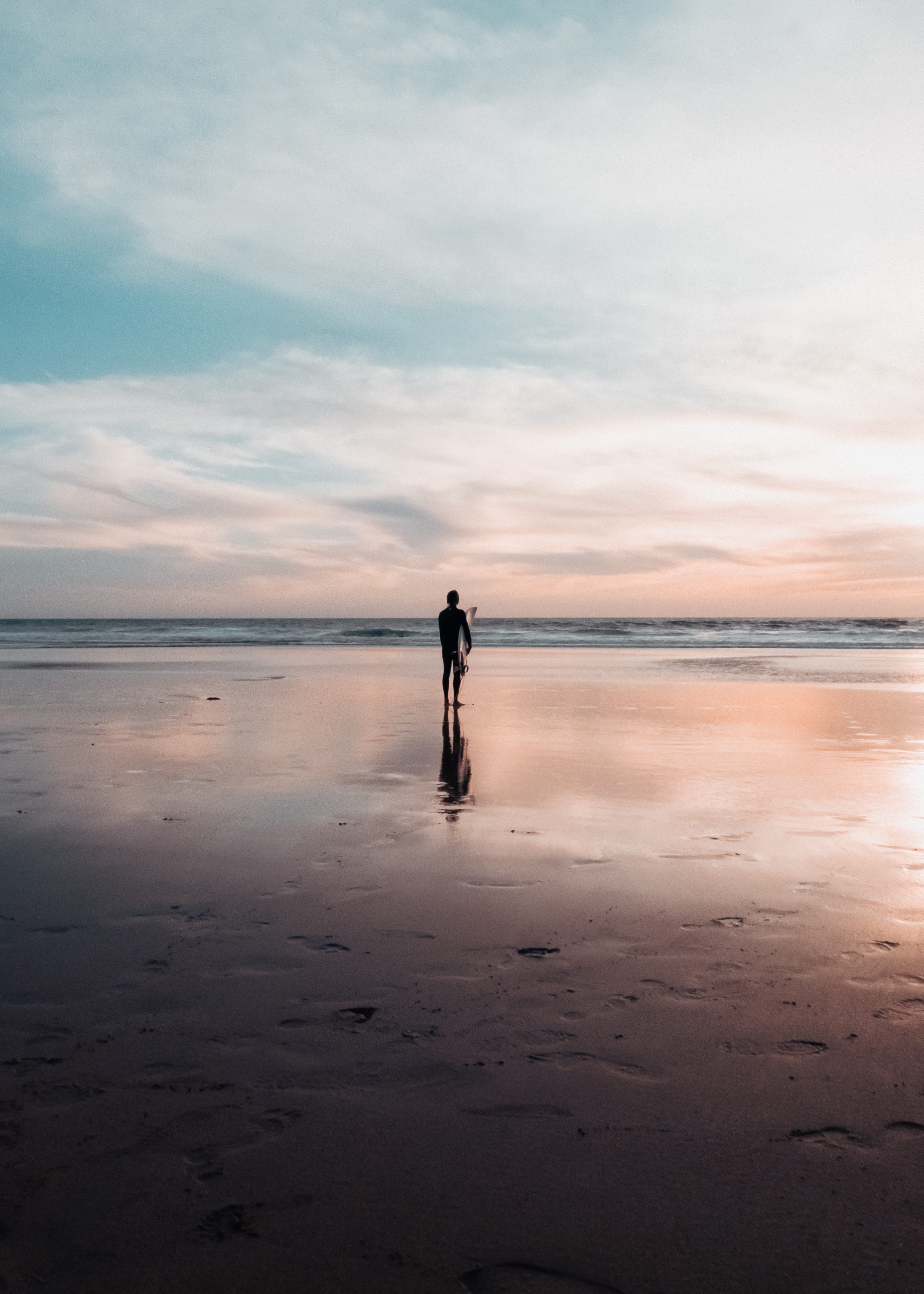 Silhouette of a surfer standing on a beach in Algarve, Portugal at sunset with reflections on the wet sand.