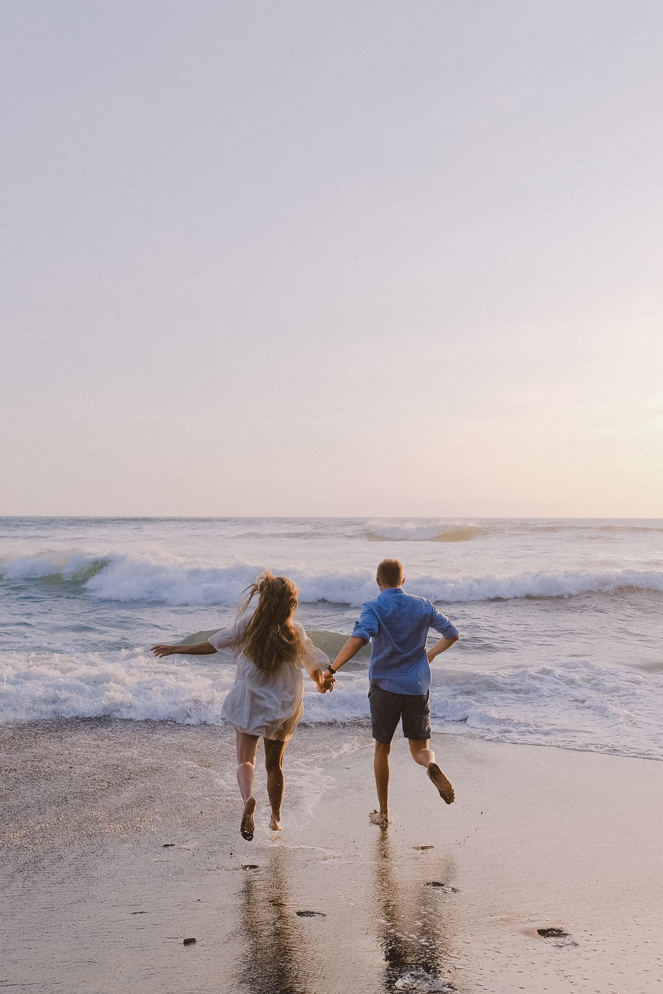 Two people running hand in hand on a beach, with waves gently breaking at their feet.