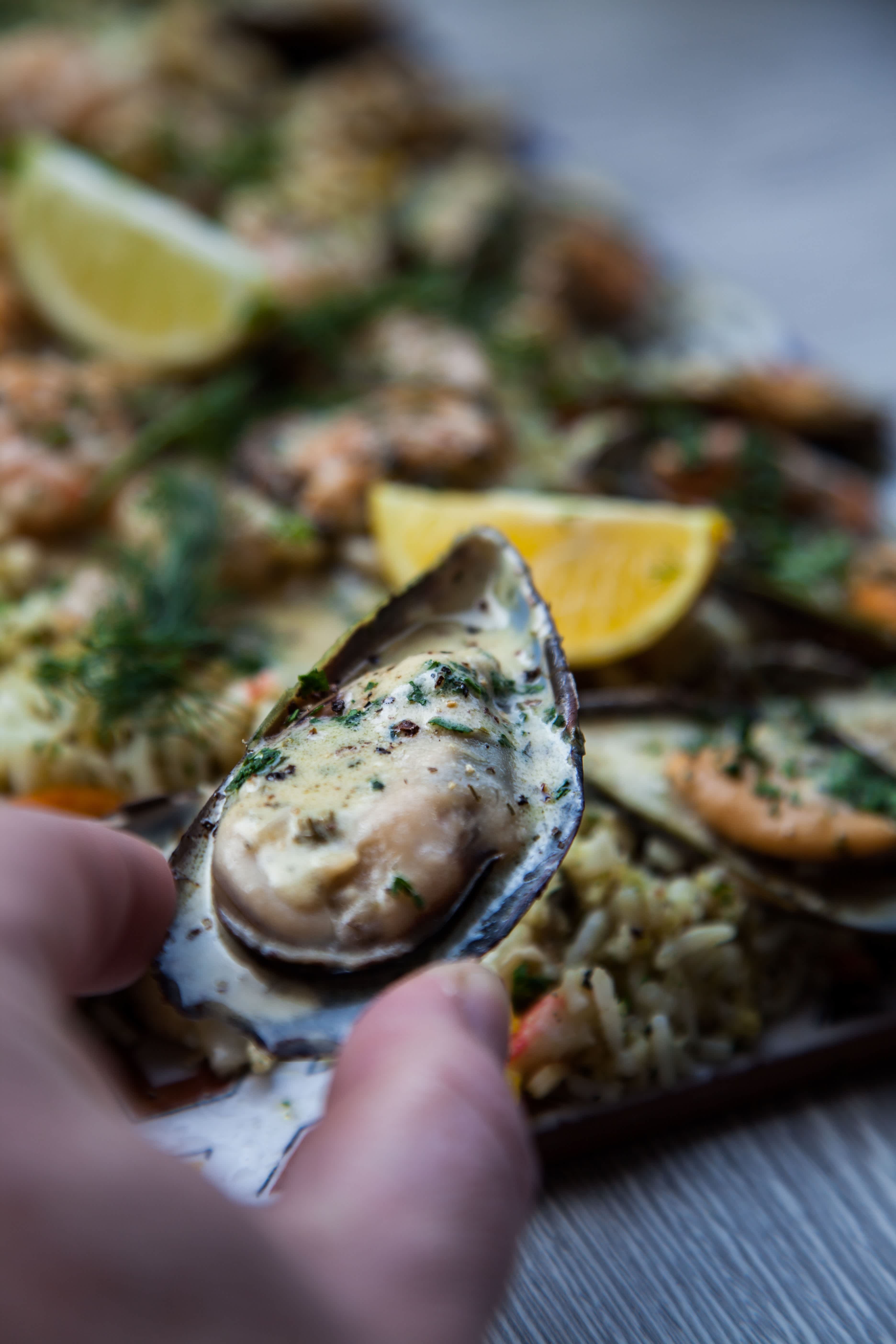 Close-up of a hand holding a grilled mussel over a platter of assorted shellfish with lemon garnish.