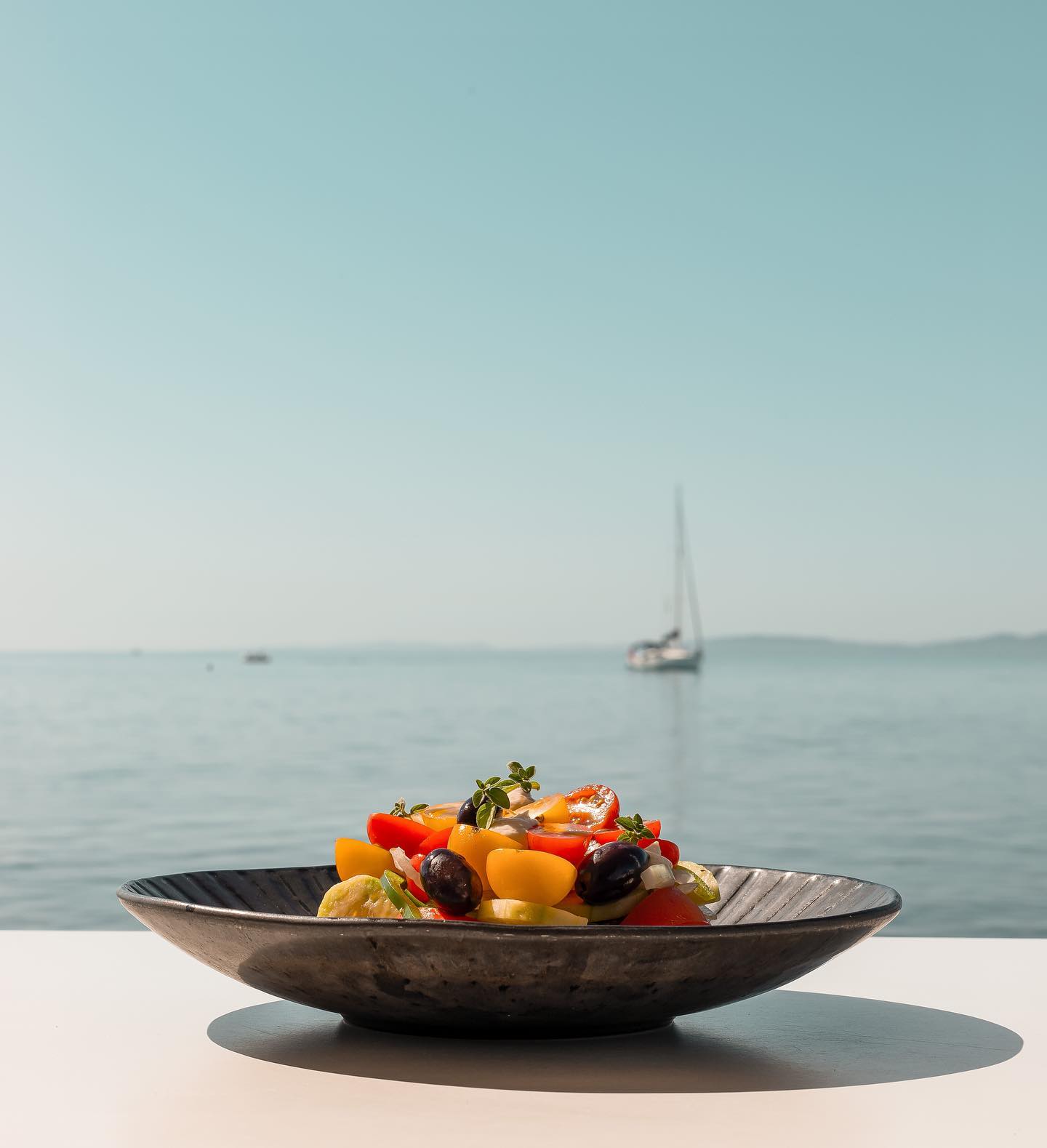 A bowl of fresh salad with a view of the sea and a sailboat in Corfu.