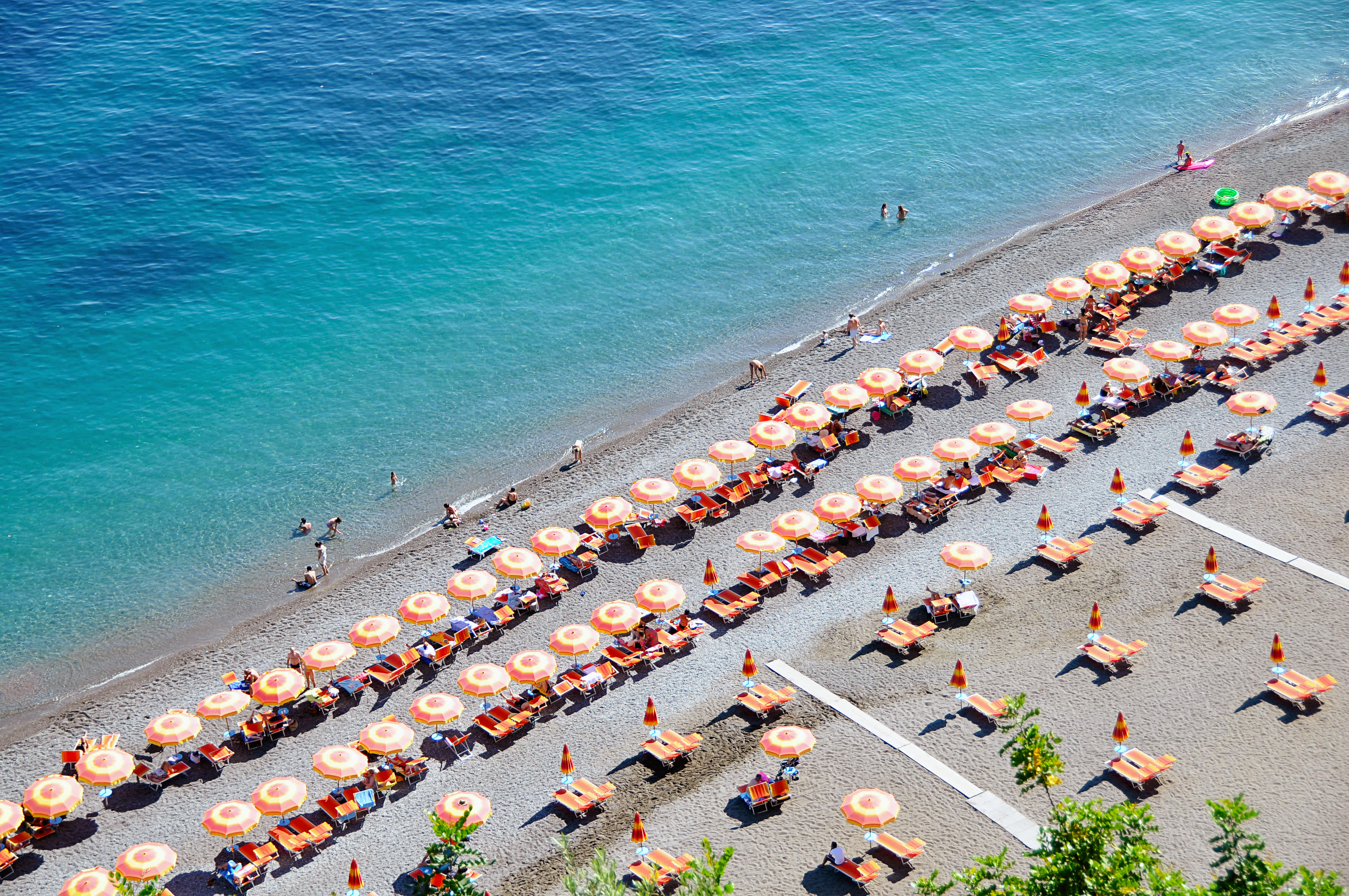 Aerial view of a beach on the Amalfi Coast with rows of orange umbrellas and sun loungers along the shoreline.