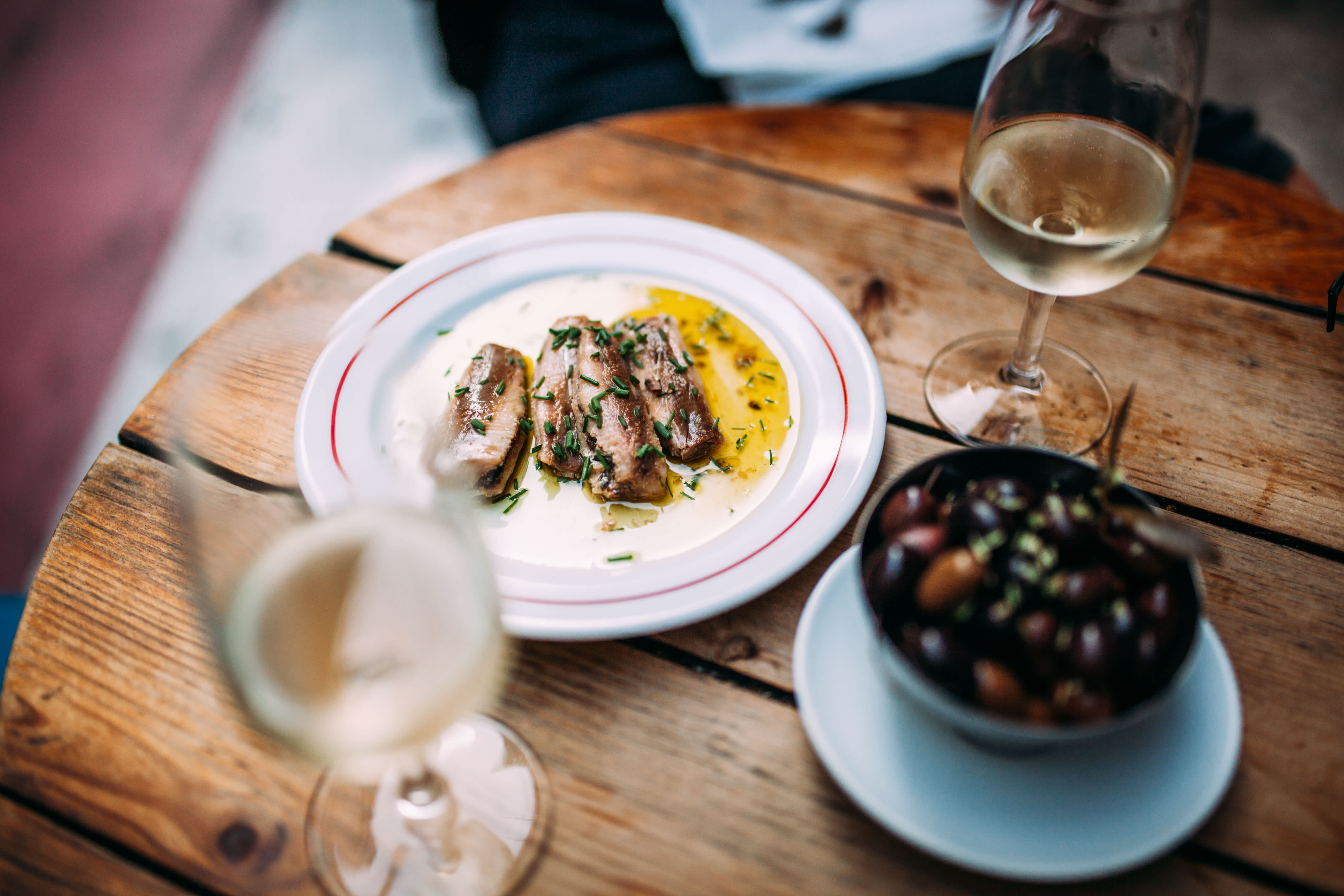 A plate of sardines with olive oil and herbs served with olives and white wine on a wooden table.