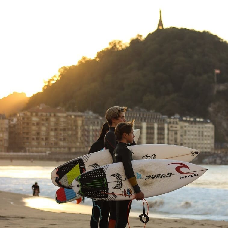 Three young surfers holding their boards and looking at the waves on a San Sebastian beach at sunset.