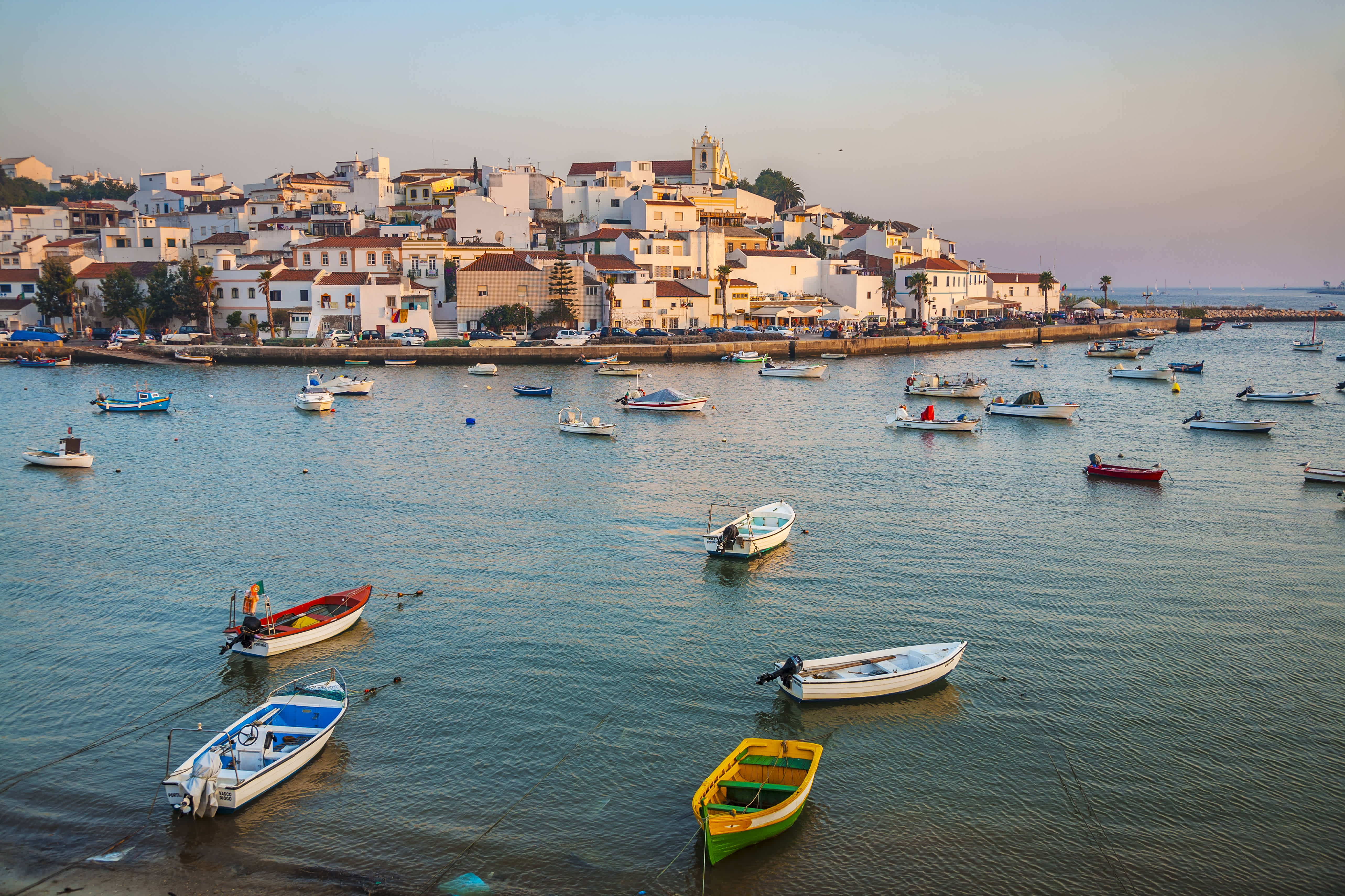 Boats floating in the harbor with a view of a coastal village in Algarve, Portugal at sunset.