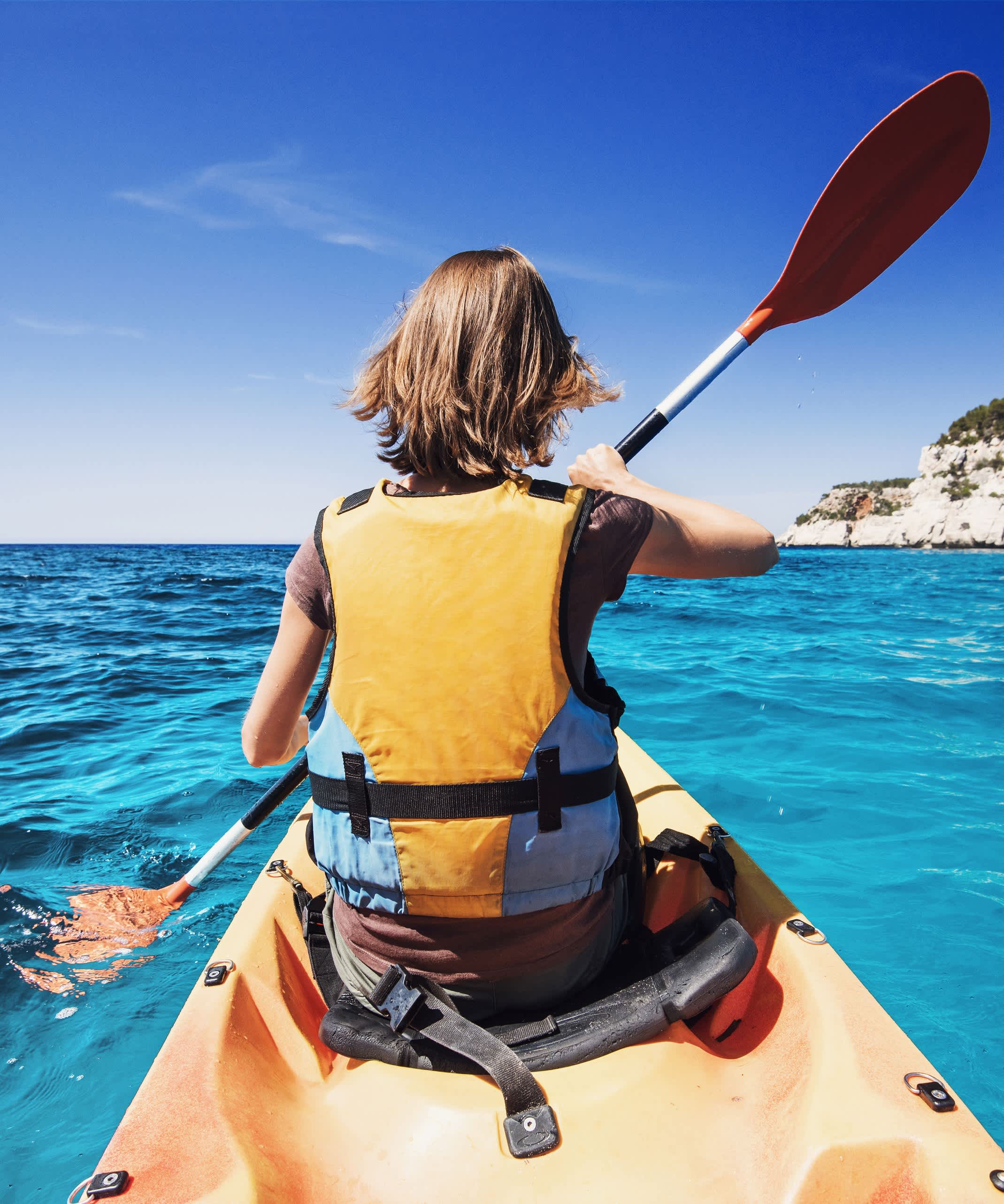 Person kayaking in clear blue waters, with a clear sky and distant rock formations.