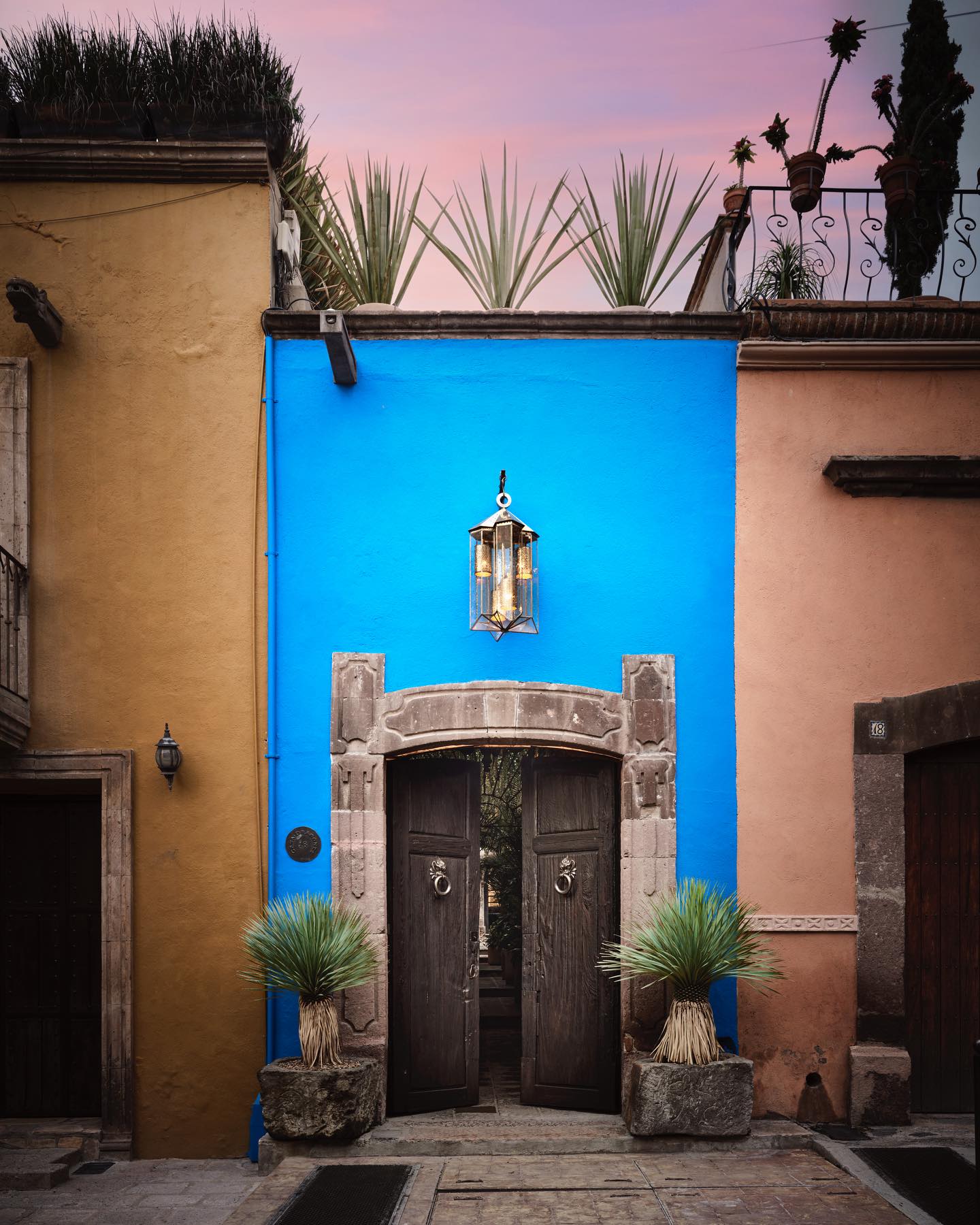 Vibrant blue entrance of a building with wooden doors and potted plants on either side, set against a sunset sky.