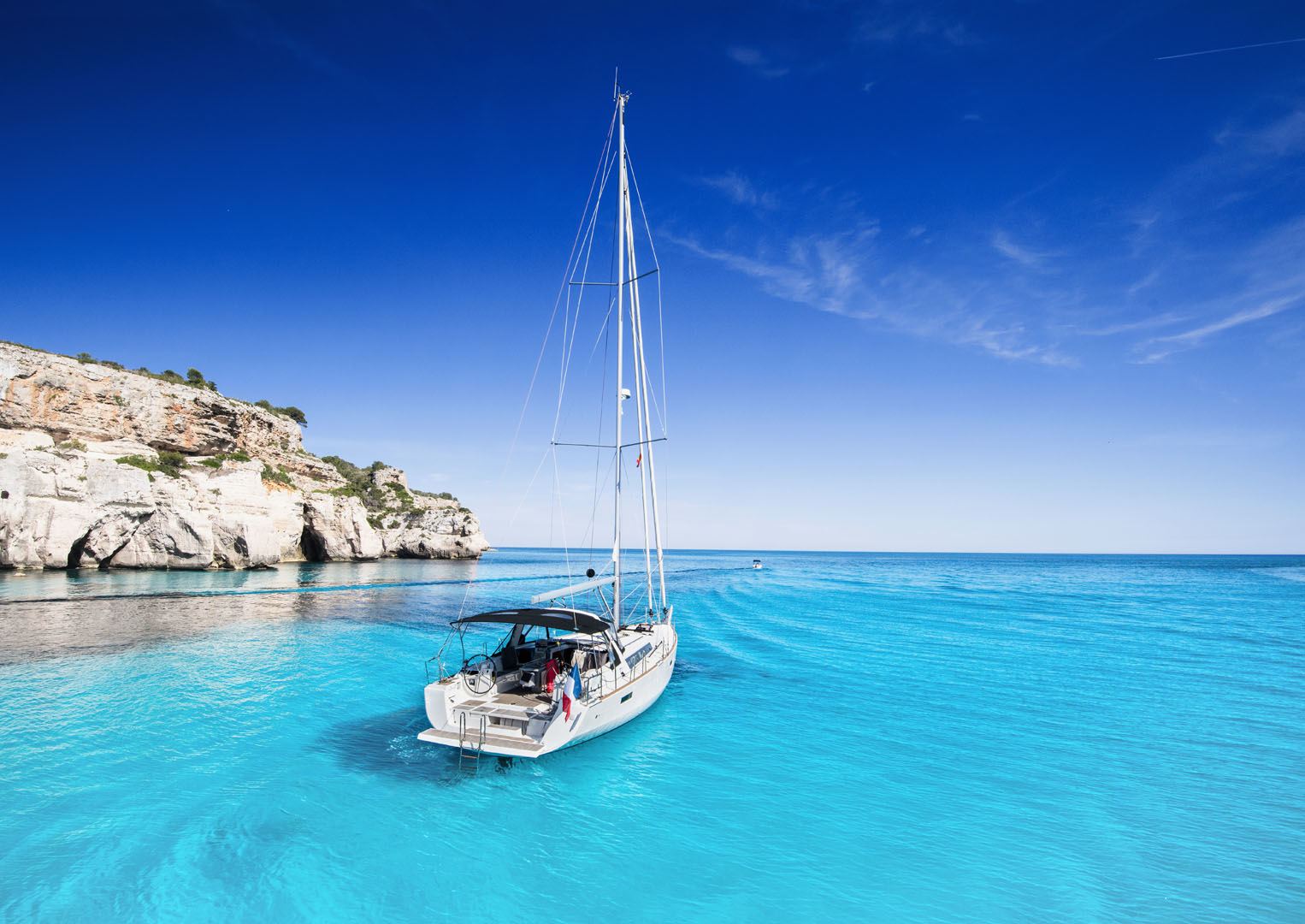 Sailboat anchored in crystal-clear turquoise waters near rocky cliffs in the Mediterranean Sea.