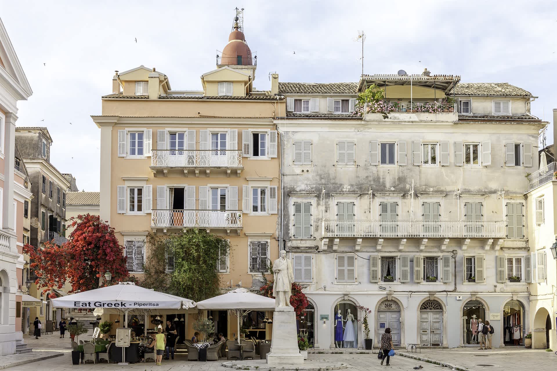 Charming square in Corfu with pastel-colored buildings, outdoor café, and a statue in the center.