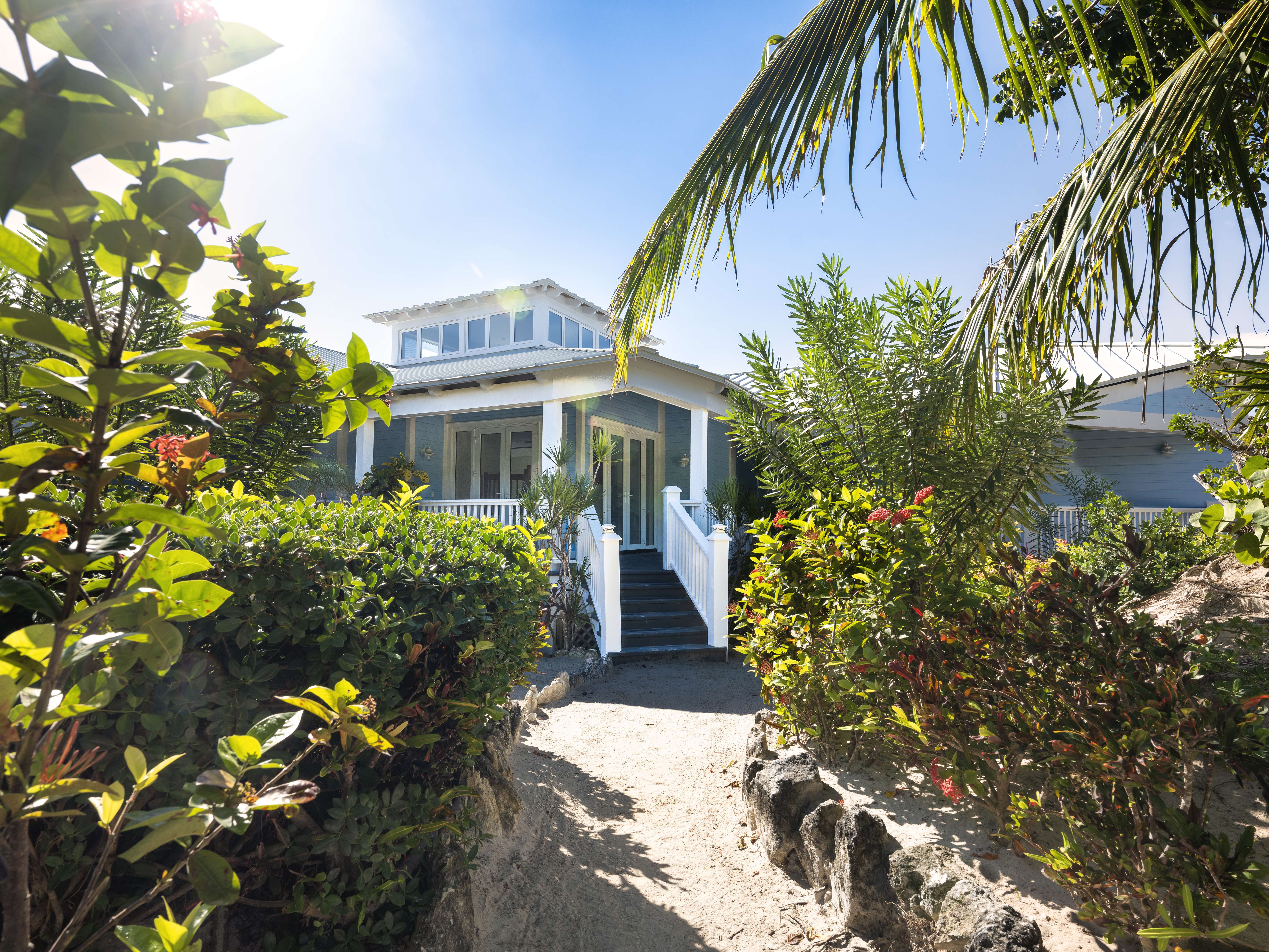 Entrance of an Exclusive Resorts property at The Abaco Club, nestled among lush tropical foliage, with a sandy path leading to a sunlit porch.