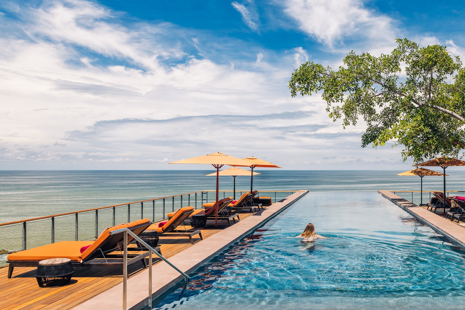Infinity pool with a swimmer, surrounded by orange-cushioned lounge chairs and umbrellas, overlooking the ocean.