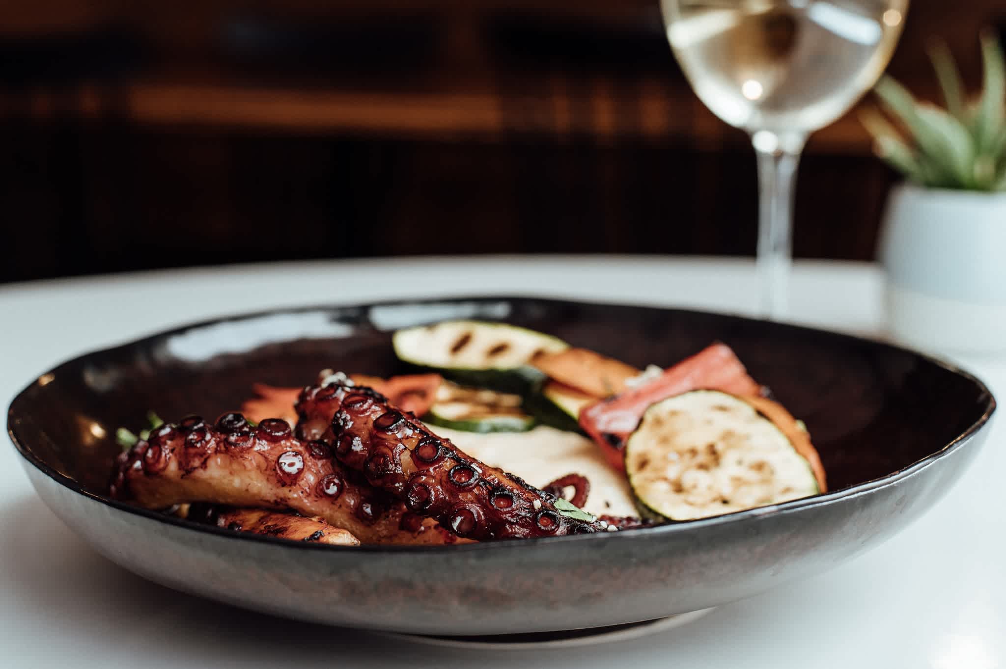 A plate of grilled octopus with vegetables served with a glass of white wine.