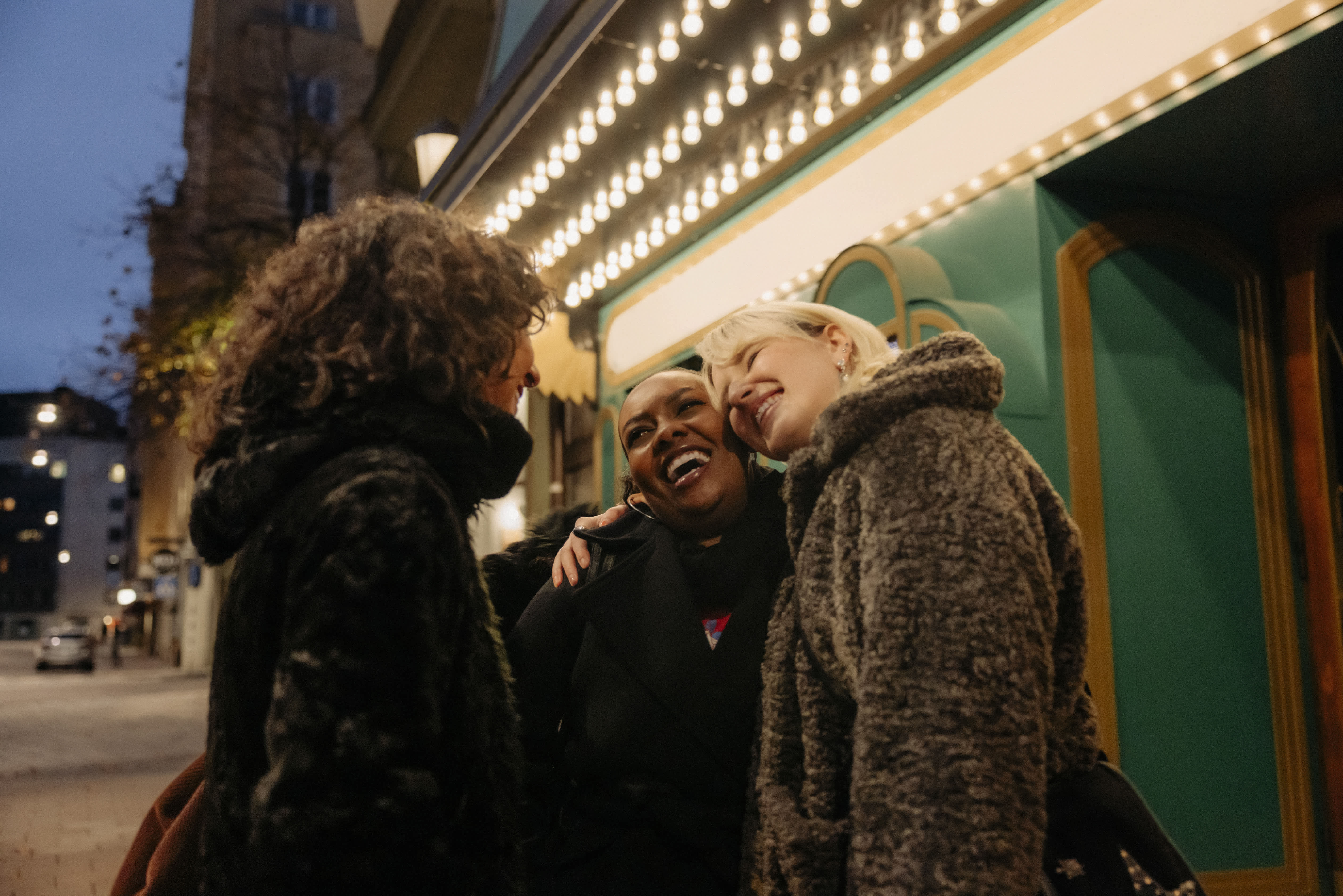 Three friends laughing and hugging outside a brightly lit building in the evening, dressed warmly in winter coats.