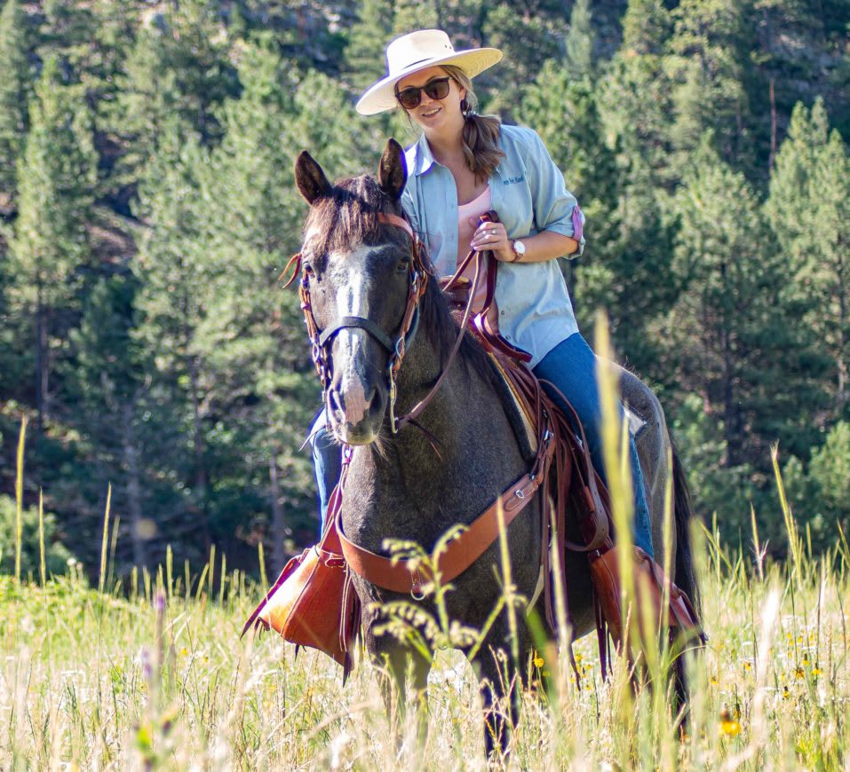Woman riding a horse through a grassy meadow at Vermejo Park Ranch, wearing a hat and sunglasses.