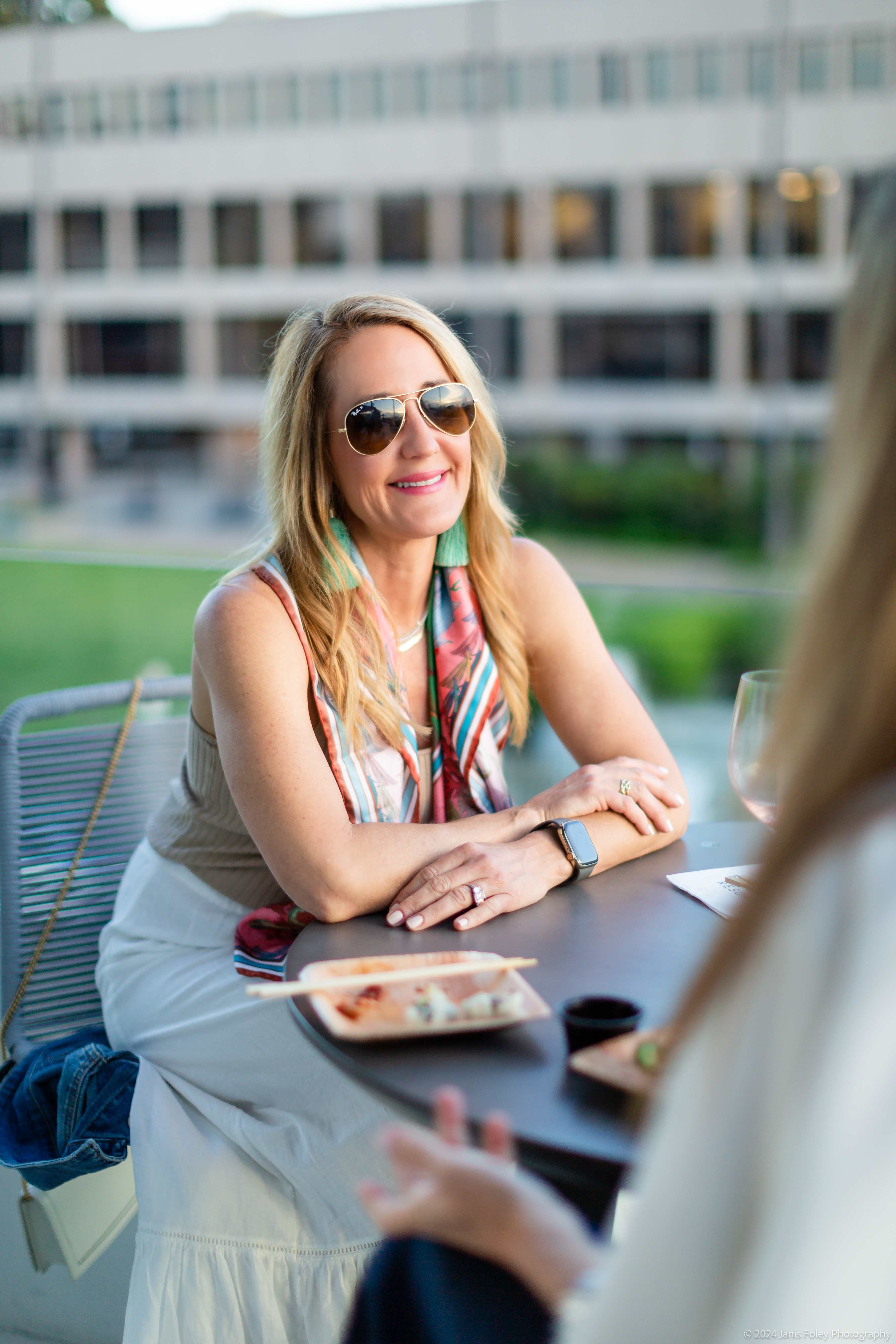 A woman sitting at a table outdoors, talking with another woman and drinking a glass of wine