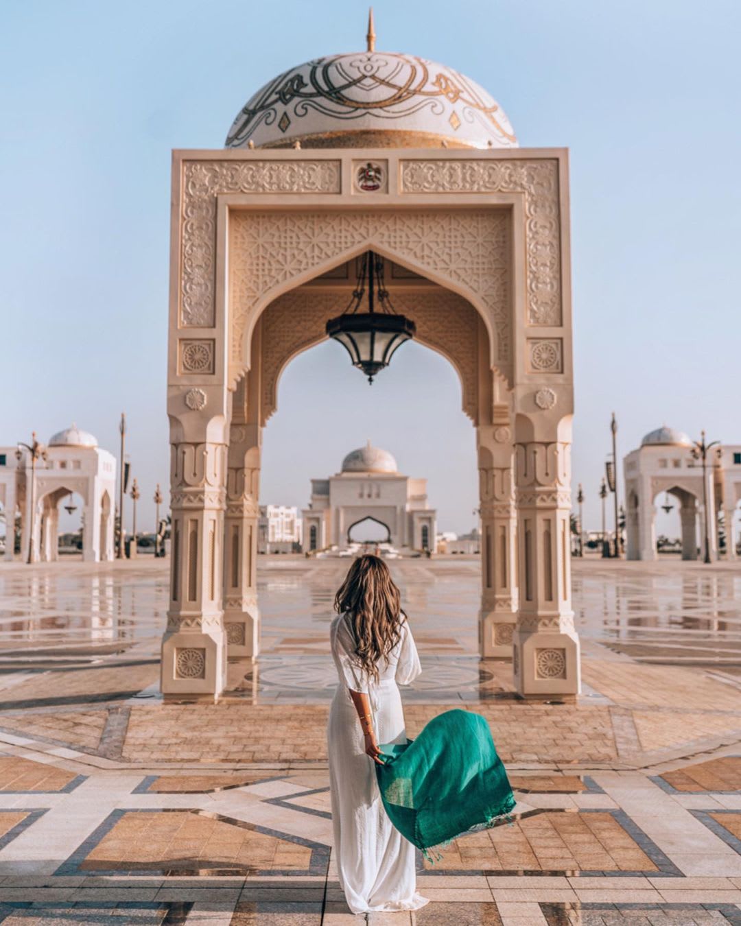A woman walking toward an ornate archway at a grand architectural site in Dubai, wearing a white dress and holding a green scarf.