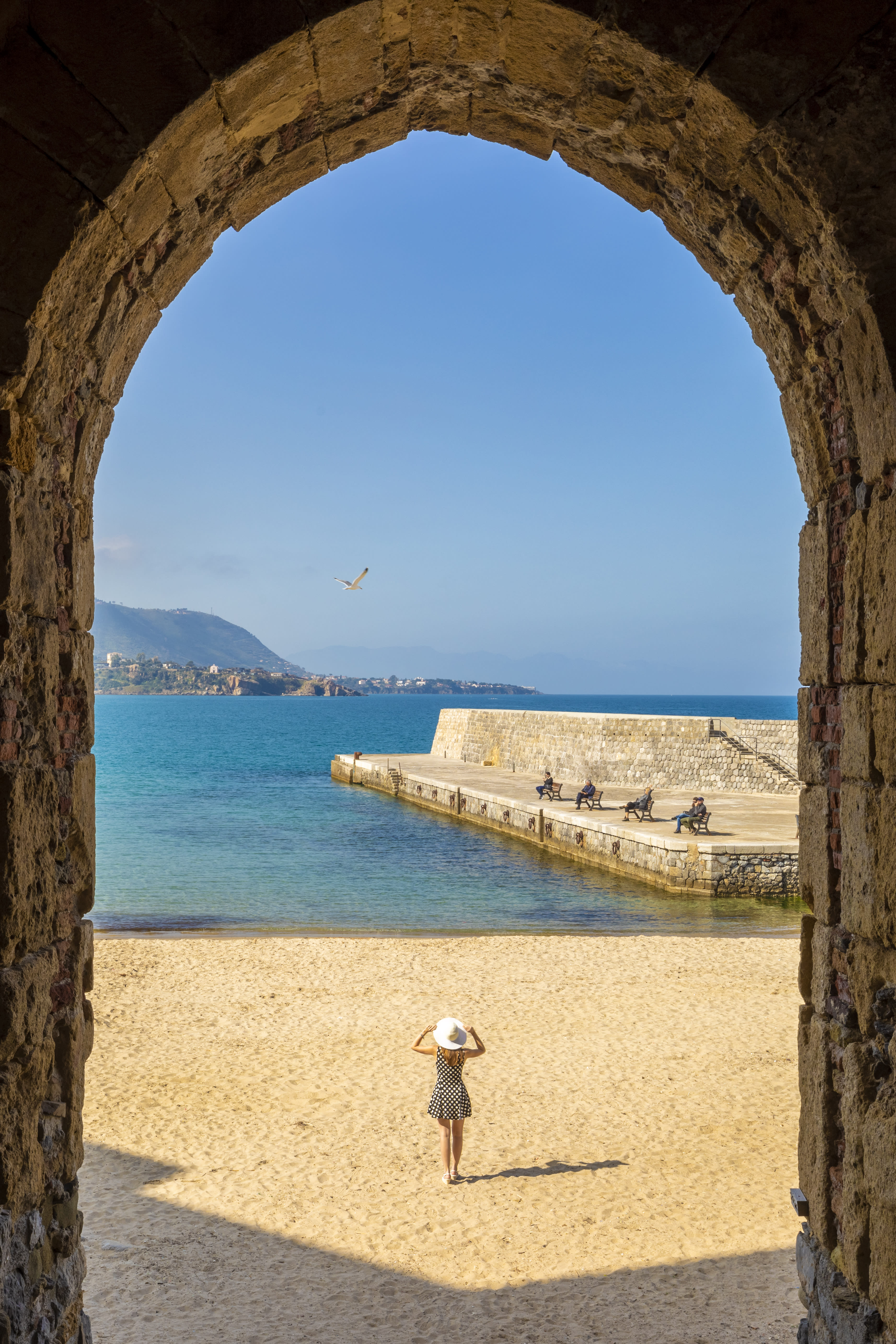 View through a stone archway of a woman standing on a sandy beach, overlooking the blue ocean and distant mountains.
