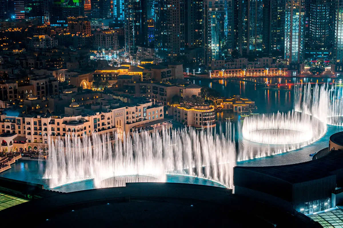 Night view of Dubai Fountain with illuminated water jets surrounded by city buildings.