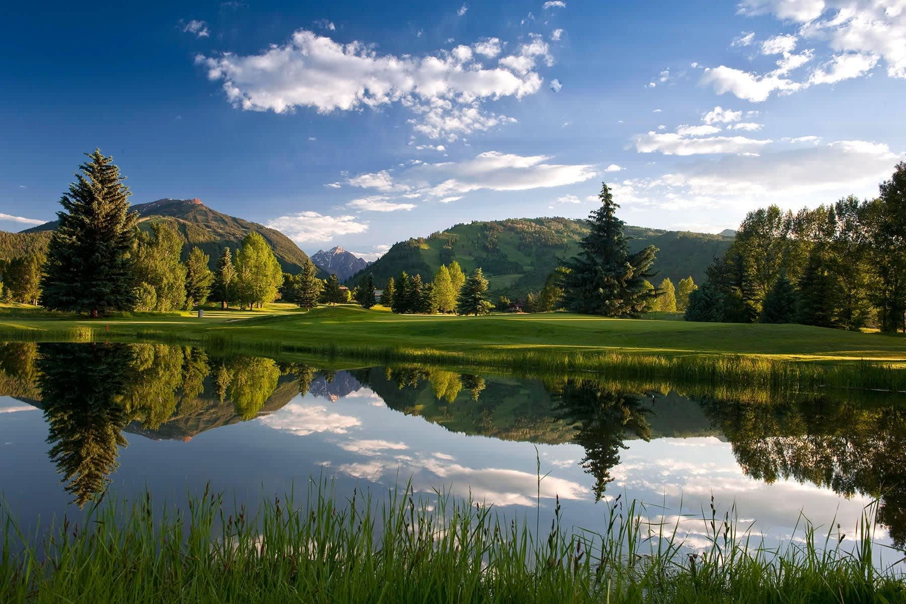Serene view of Aspen Golf Club with lush green fairways, tall trees, and a tranquil pond reflecting the surrounding mountains.