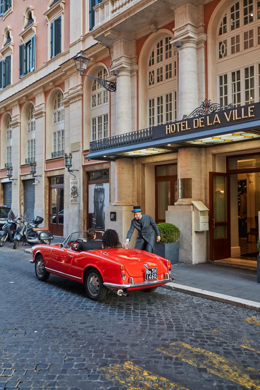 Valet assisting a couple in a red convertible outside Hotel de la Ville, with elegant architecture and cobblestone streets.