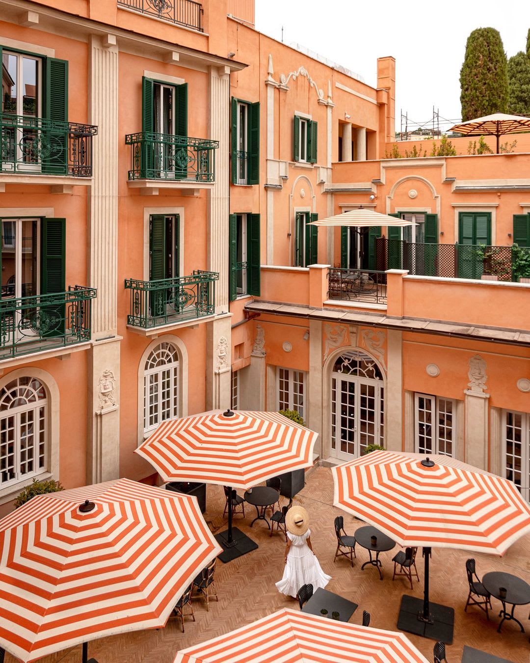 Elegant courtyard in Rome with striped umbrellas and a woman in a white dress and hat.