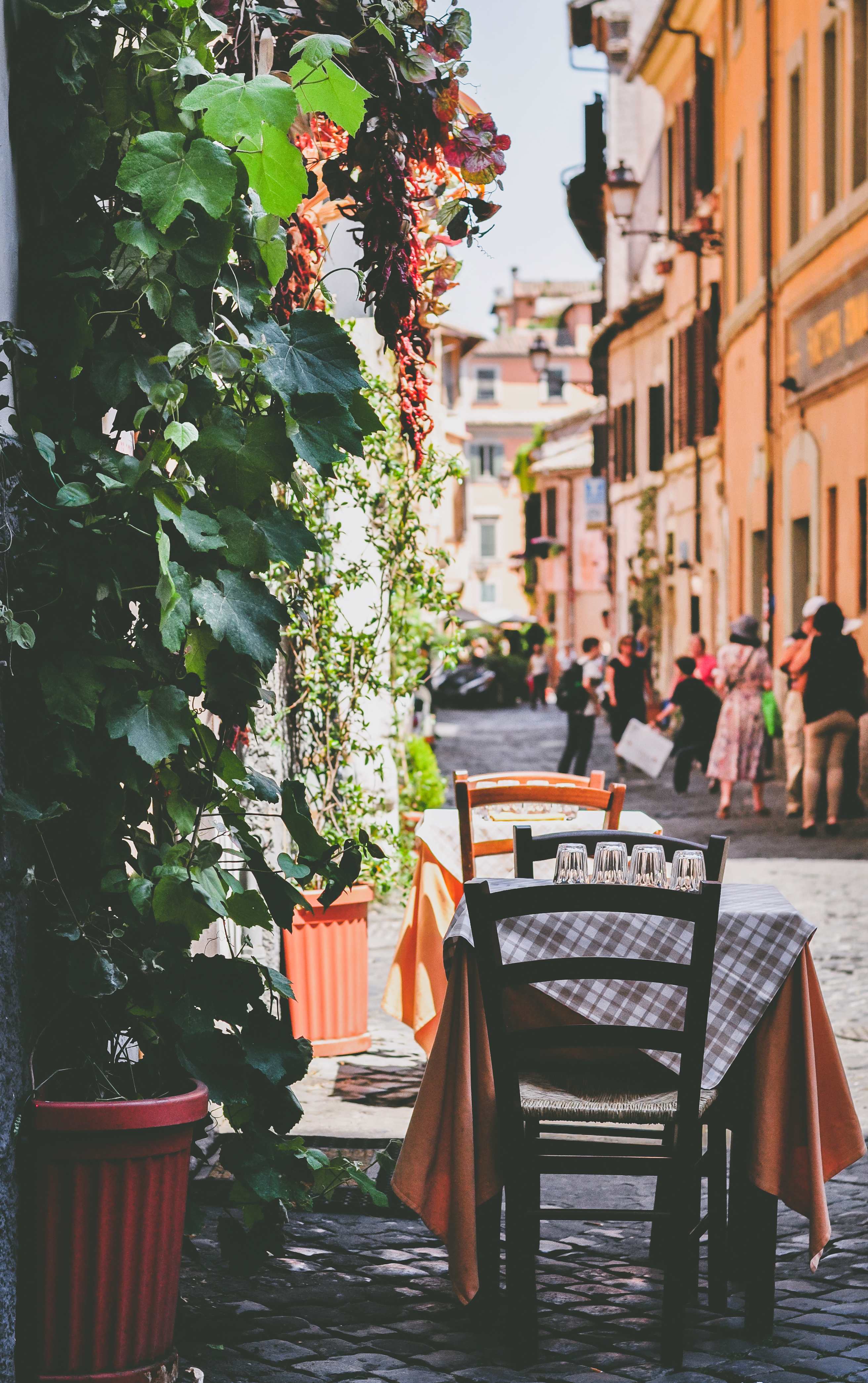 A quaint outdoor café table in Rome, adorned with a checkered tablecloth and glasses, surrounded by lush greenery and colorful buildings.