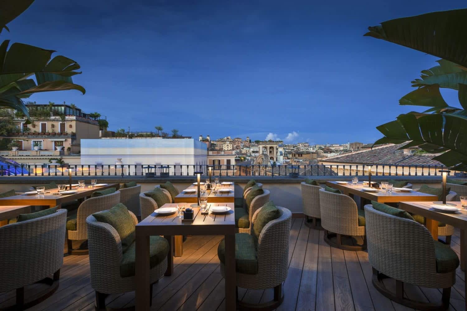 Outdoor rooftop restaurant in Rome with a city skyline view at dusk, featuring elegantly set tables and cozy seating.