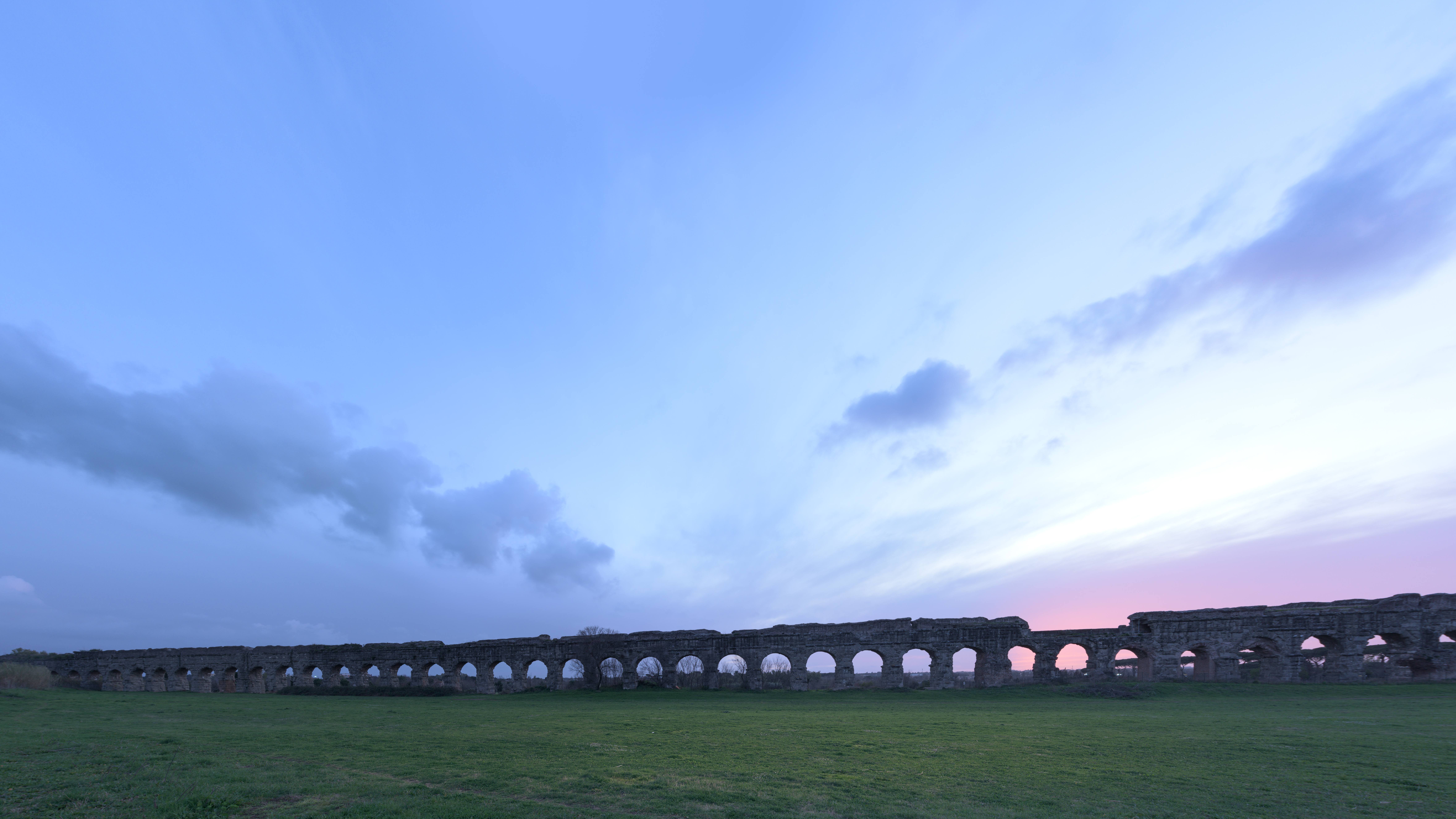 Ancient Roman ruins in a grassy field under a colorful evening sky.