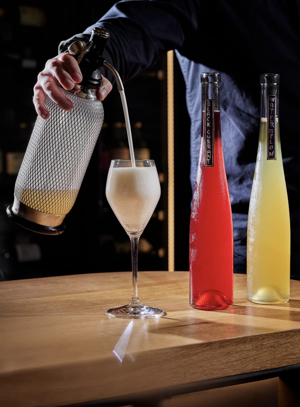 A person pouring a drink into a glass from a siphon with bottles labeled 'Black Currant' and 'Watermelon' at Degustation Restaurant in Prague