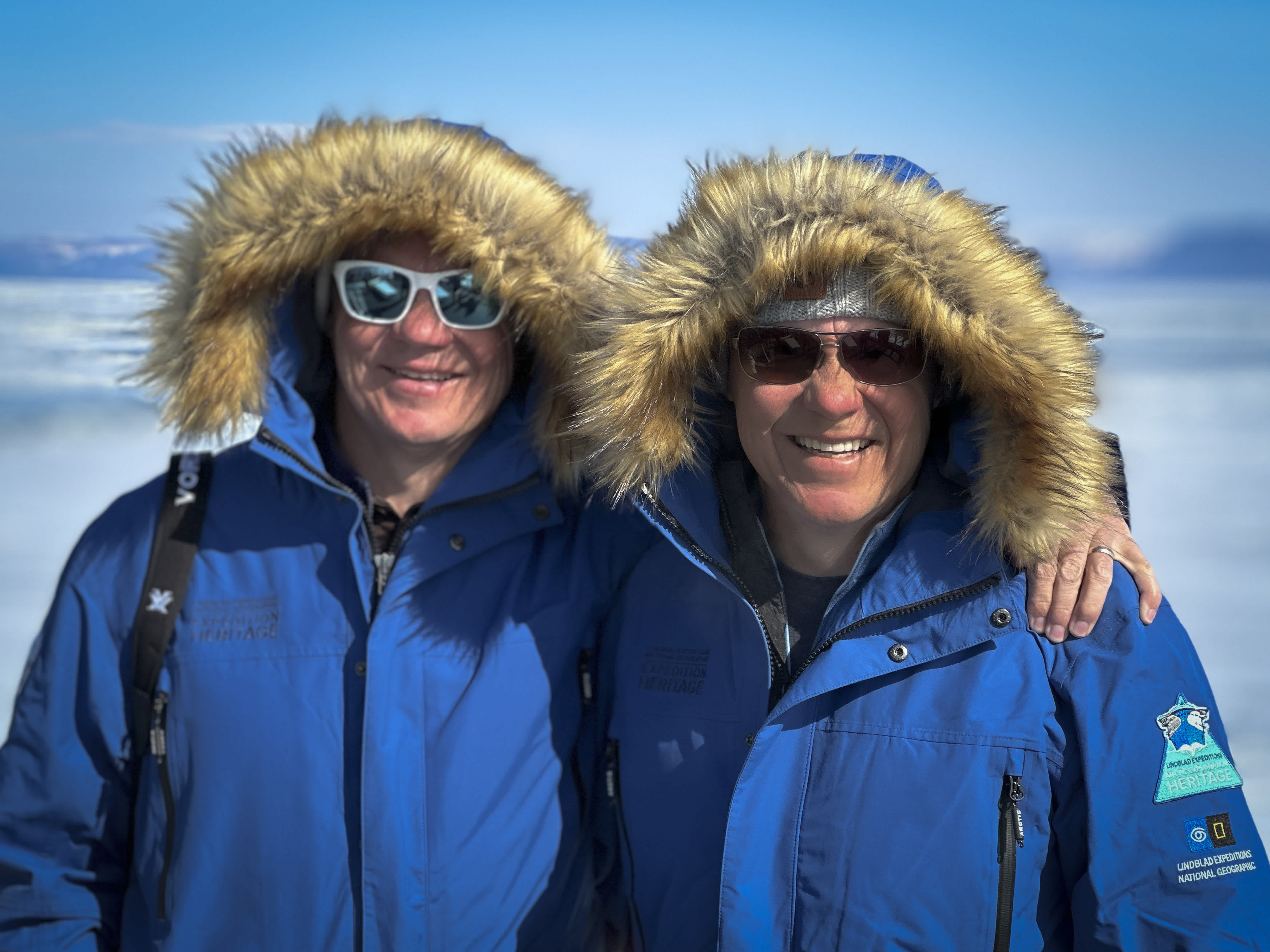 Two Exclusive Resorts' Members in blue parkas with fur-lined hoods, standing together in the Arctic.