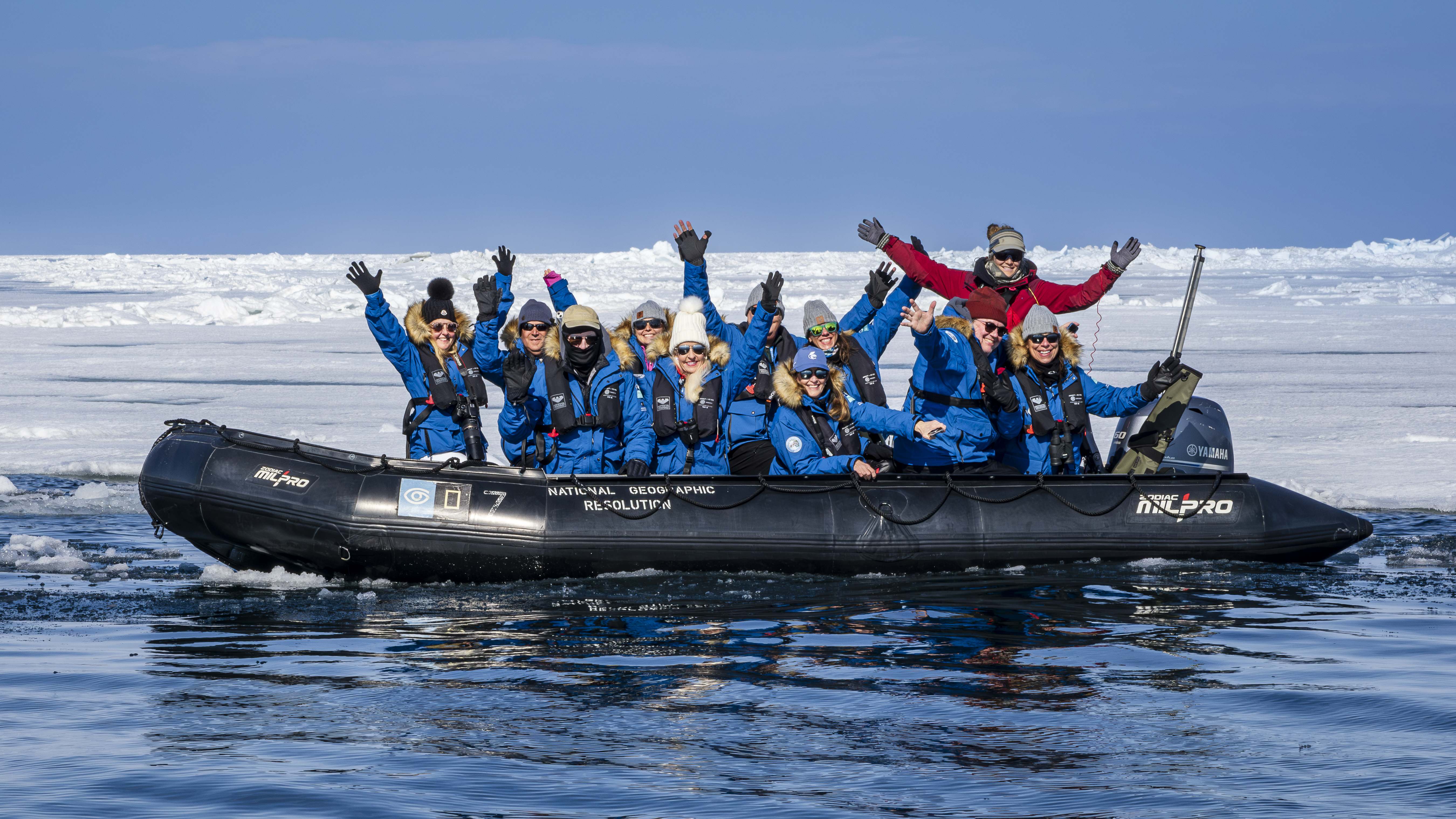 A group of Exclusive Resorts' Members in blue jackets, smiling and waving while riding in a Zodiac boat on icy Arctic waters.