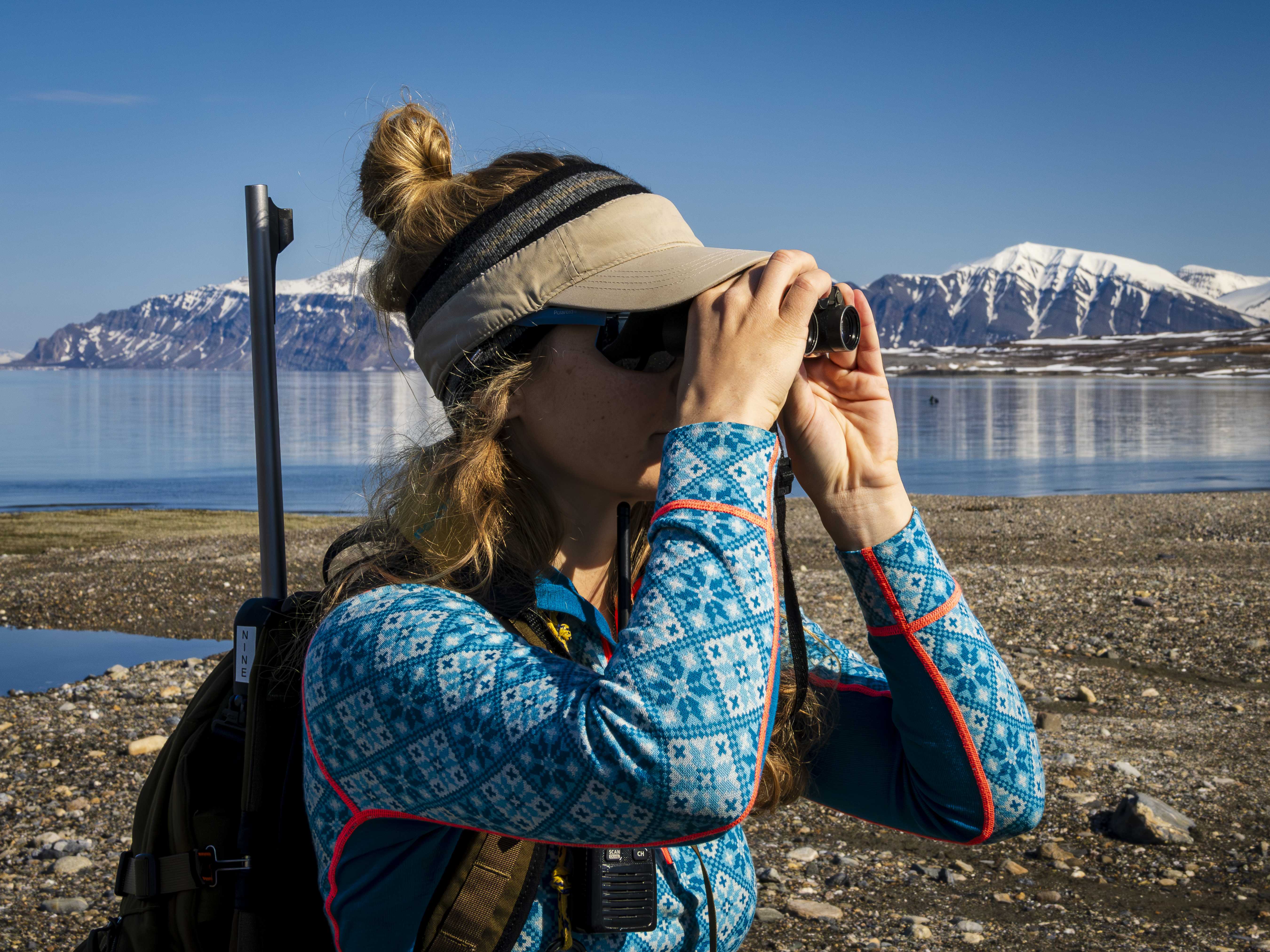 An Exclusive Resorts' Member using binoculars to observe the Arctic landscape, with mountains and calm water in the background.