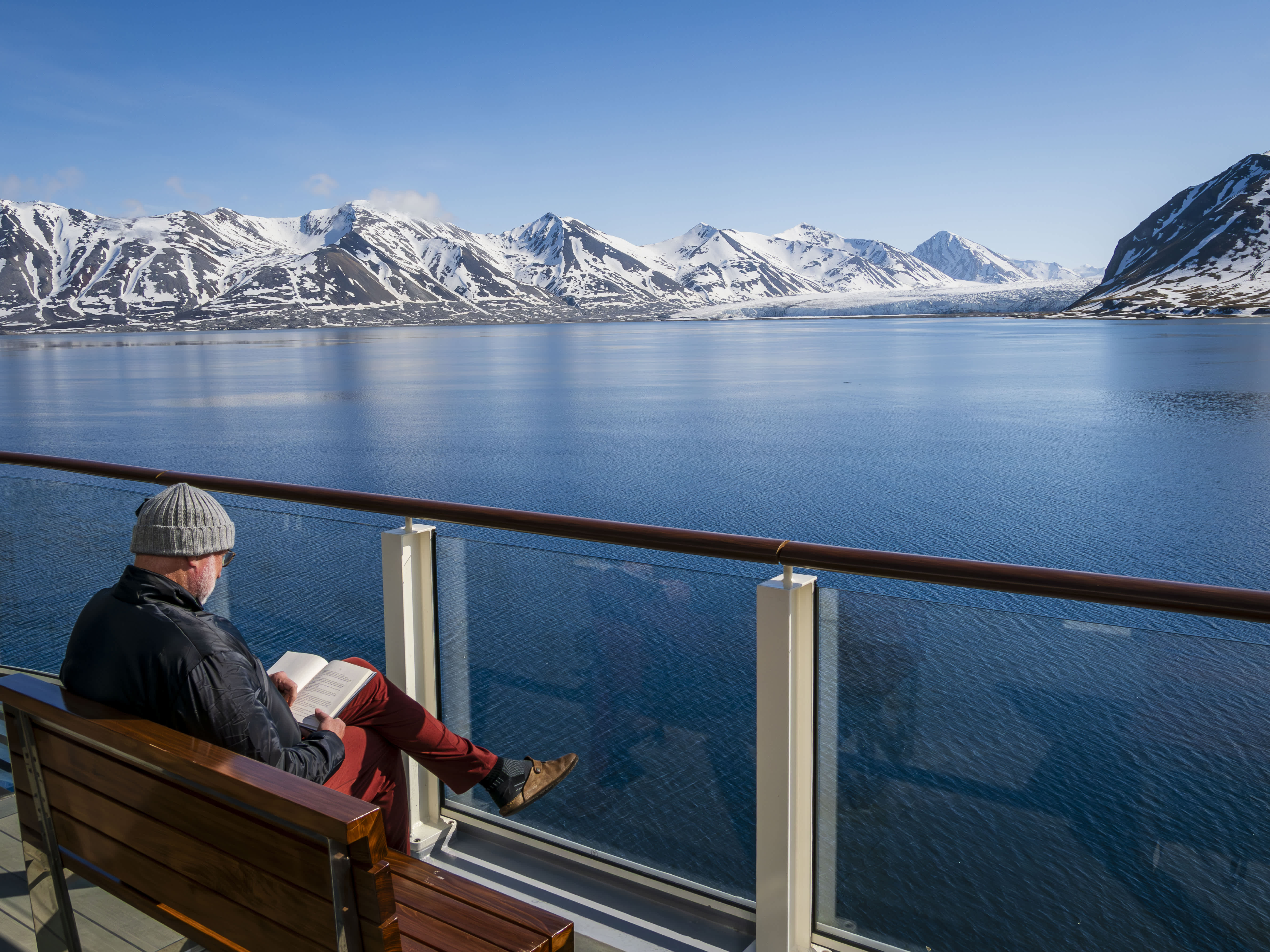 An Exclusive Resorts' Member reading a book aboard the National Geographic Resolution, overlooking the calm Arctic waters and snow-capped mountains.