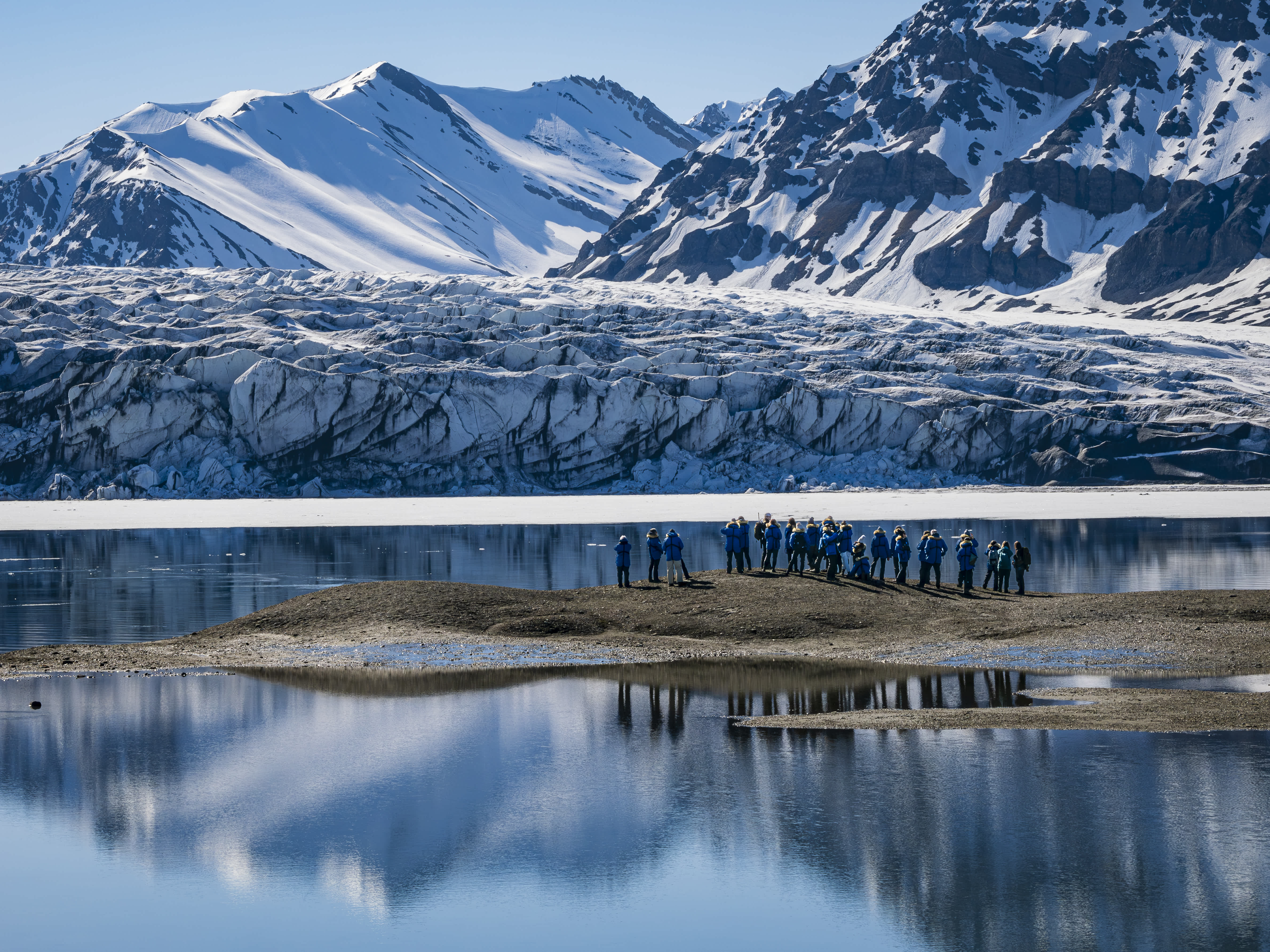 A group of people in blue jackets standing on a small island with a glacial backdrop and snowy mountains in the Arctic.
