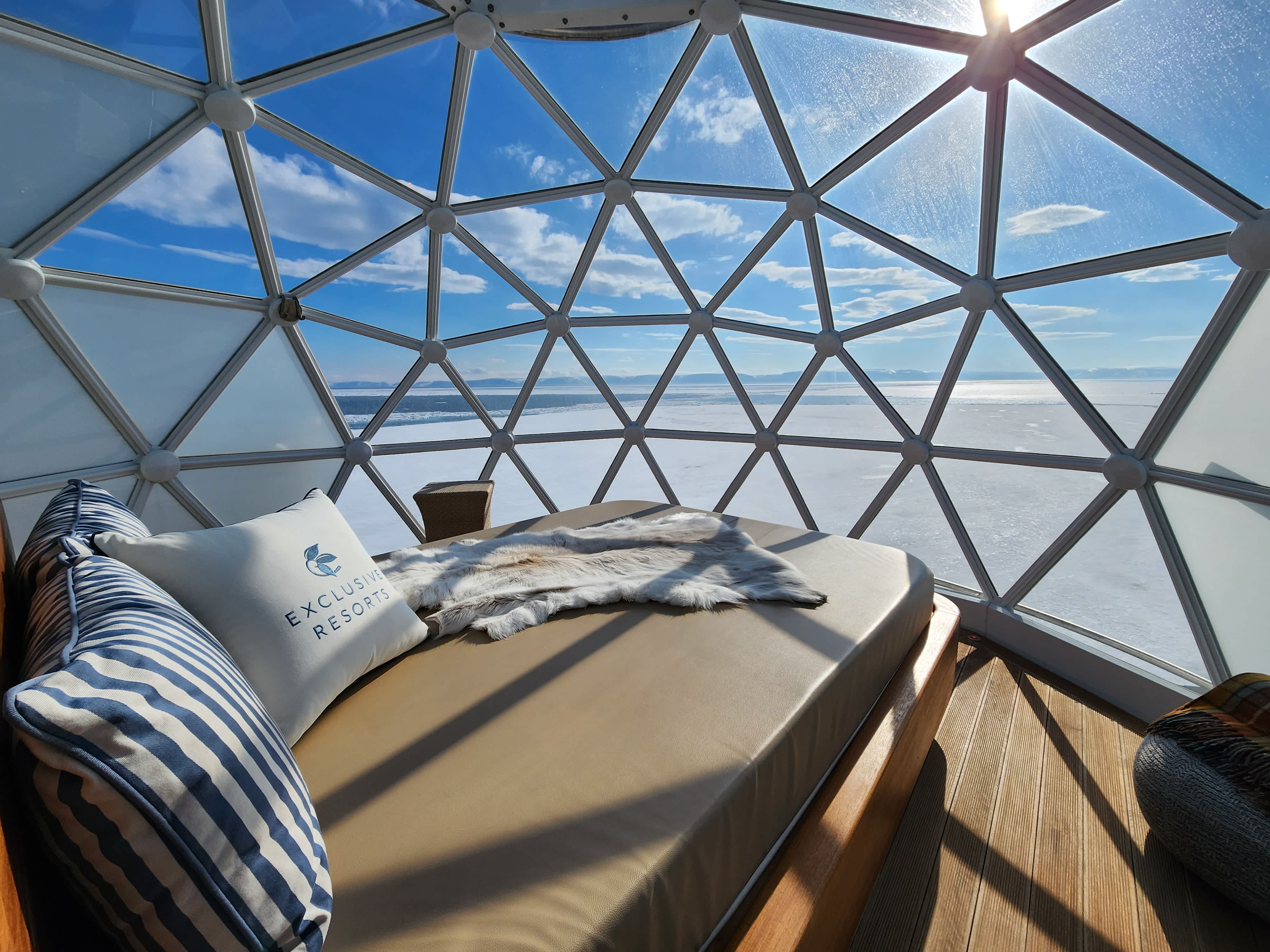 A cozy bed with an Exclusive Resorts branded pillow inside an igloo-like dome, offering a panoramic view of the snowy Arctic landscape under a bright blue sky.