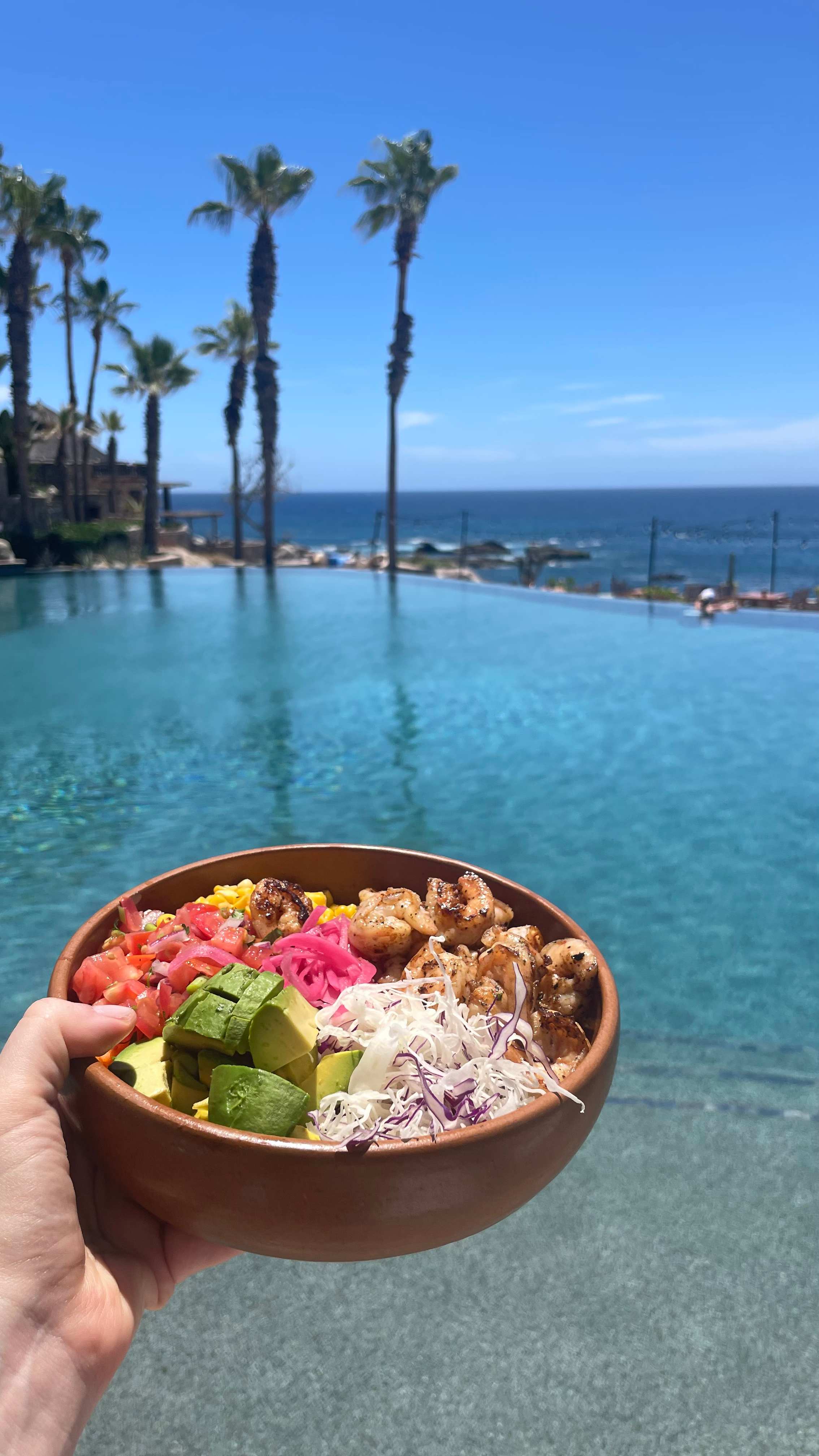 Hand holding a bowl of fresh shrimp salad with avocado, tomatoes, and cabbage in front of an infinity pool at Esperanza in Los Cabos, Mexico.