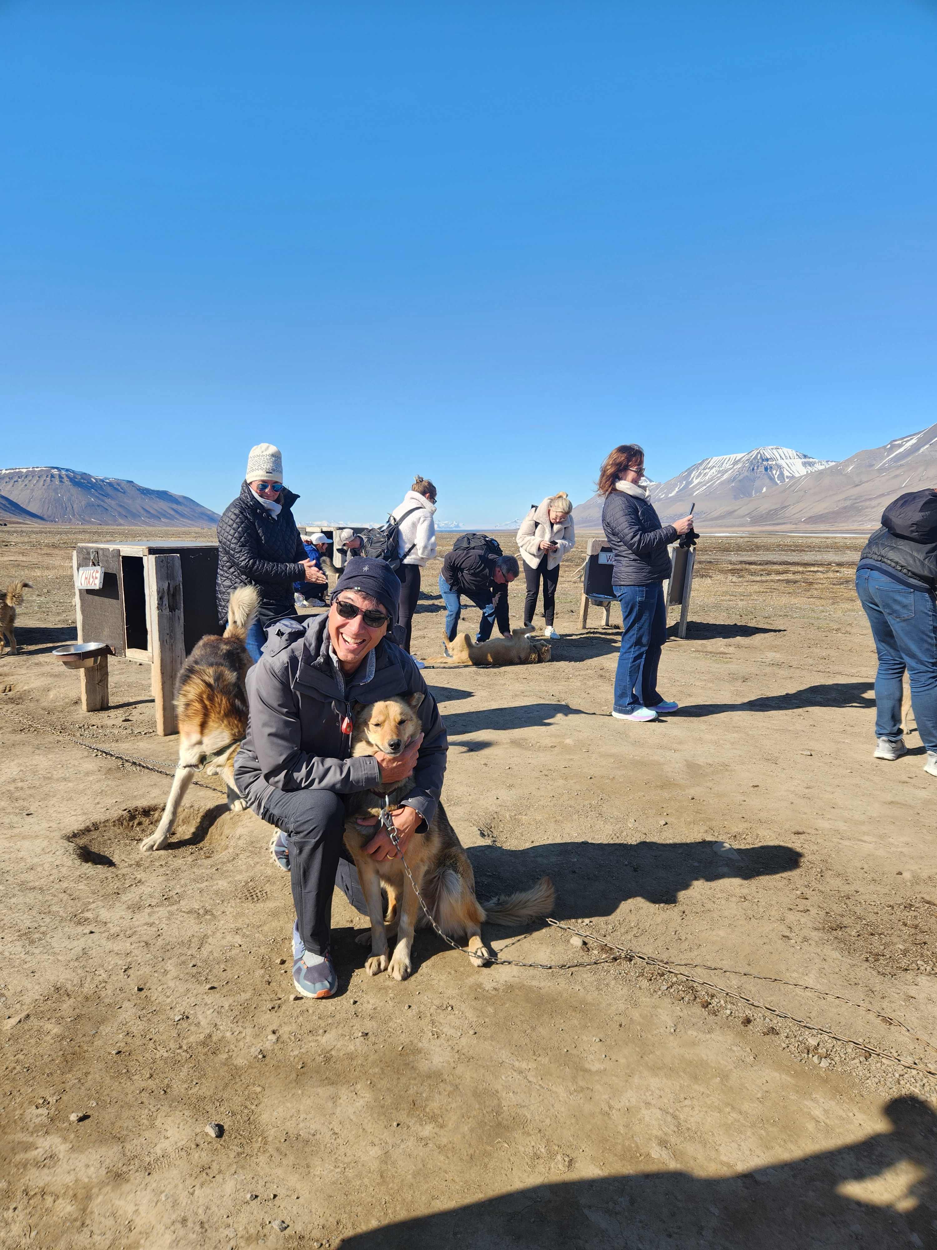 A person kneeling and hugging a brown dog at an outdoor kennel area with other people and dogs set against a backdrop of mountains and blue sky in the Arctic.