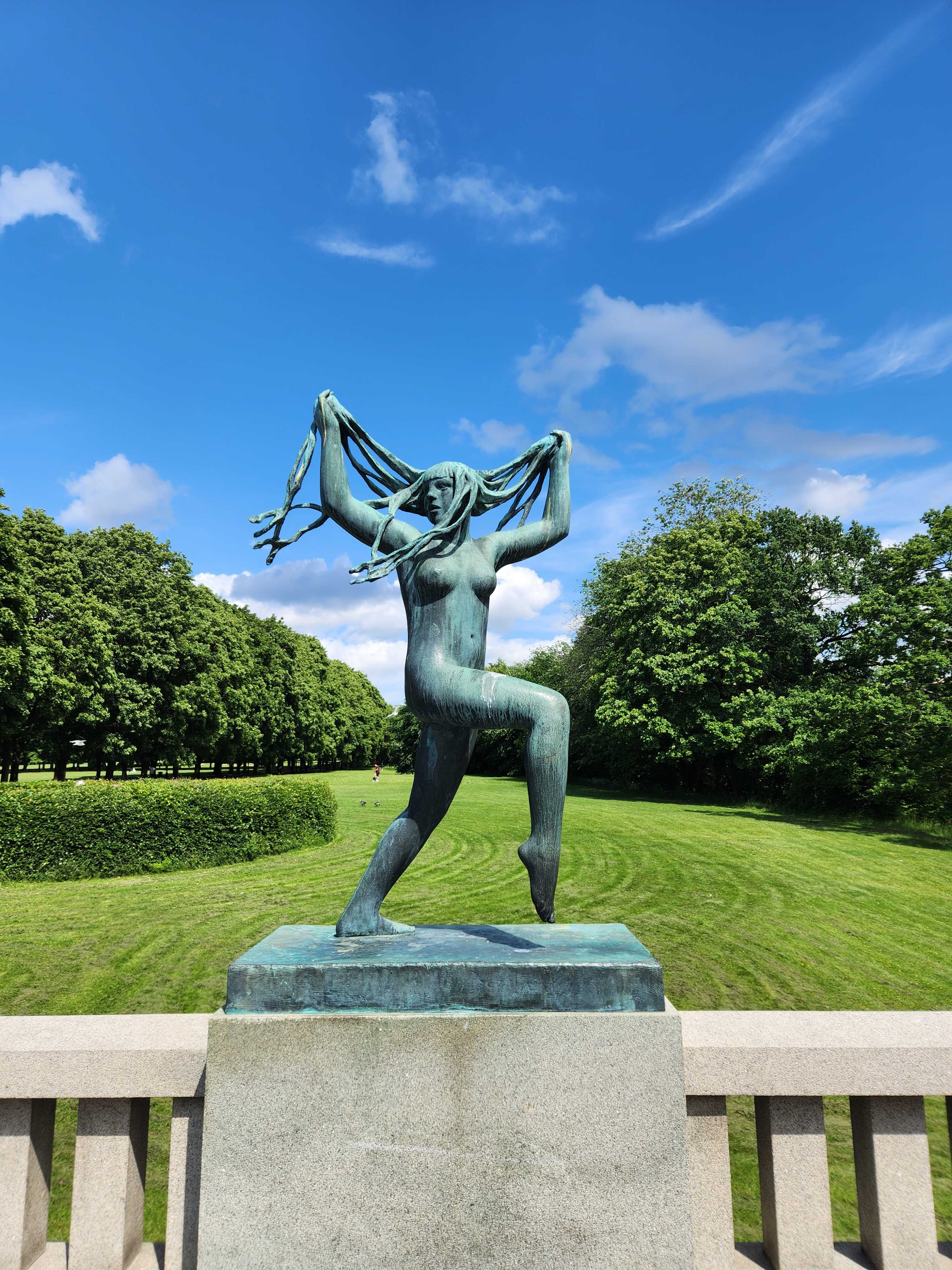 A bronze statue of a dancing woman holding her hair in the air, set against a backdrop of green trees and a blue sky in a park