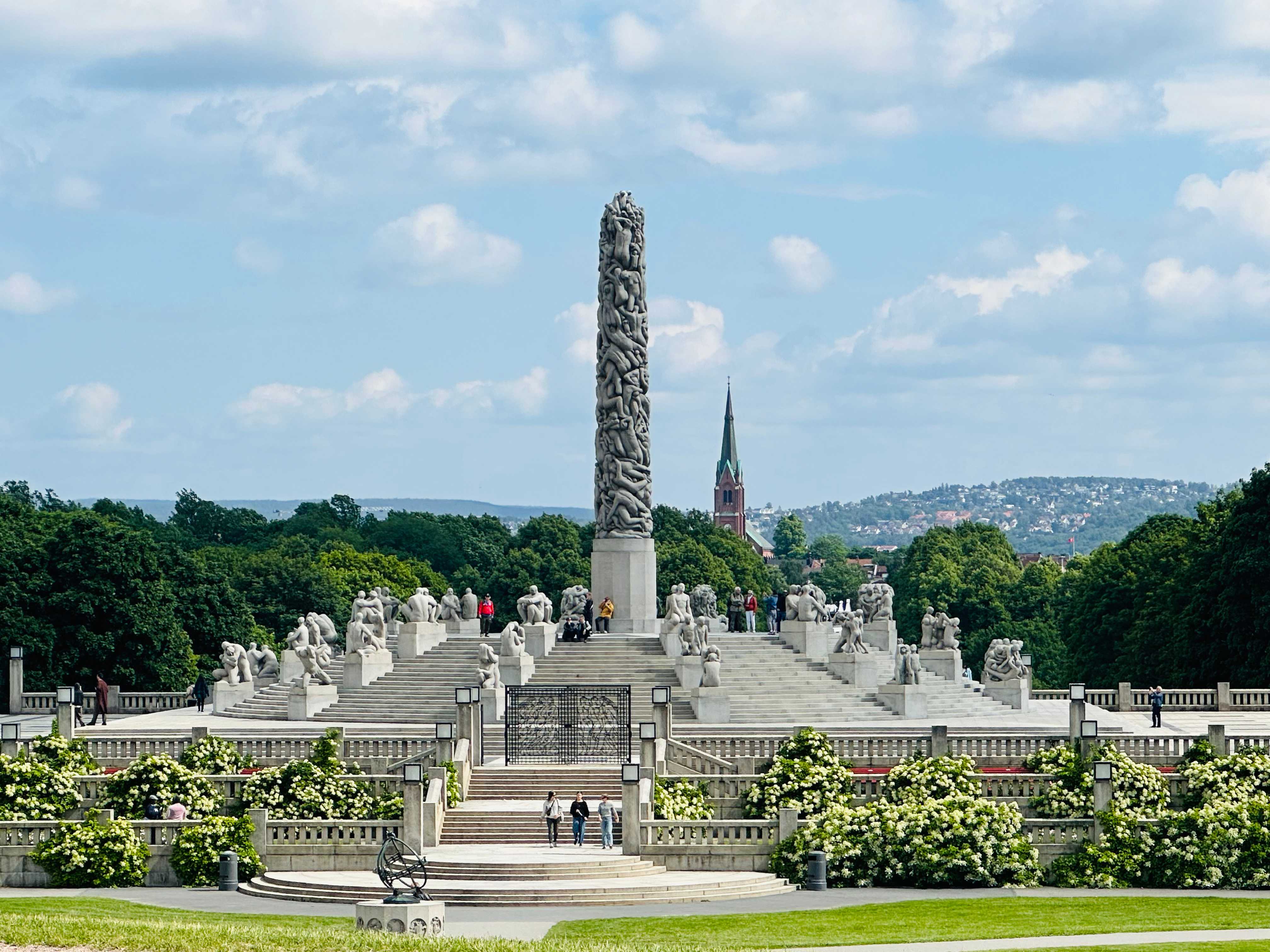 A view of the Vigeland installation in Frogner Park, Oslo, featuring the Monolith, a tall sculpture surrounded by other stone statues, with greenery and a church spire in the background.