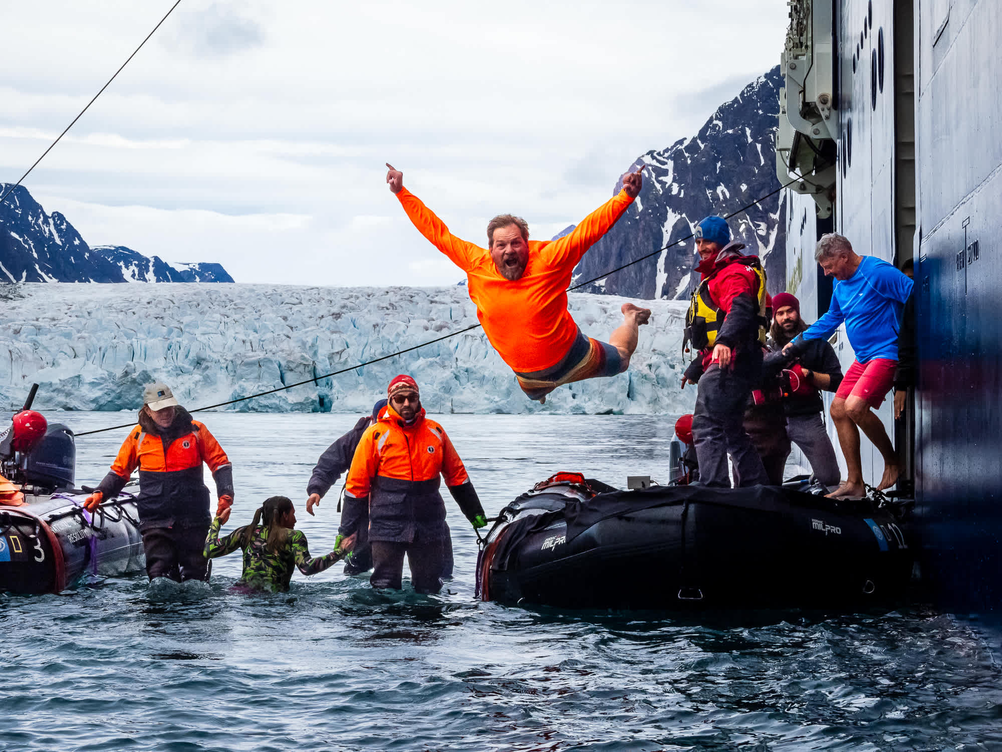 Exclusive Resorts' Members taking a plunge into icy Arctic waters from a ship, with a glacier and snow-capped mountains in the background.