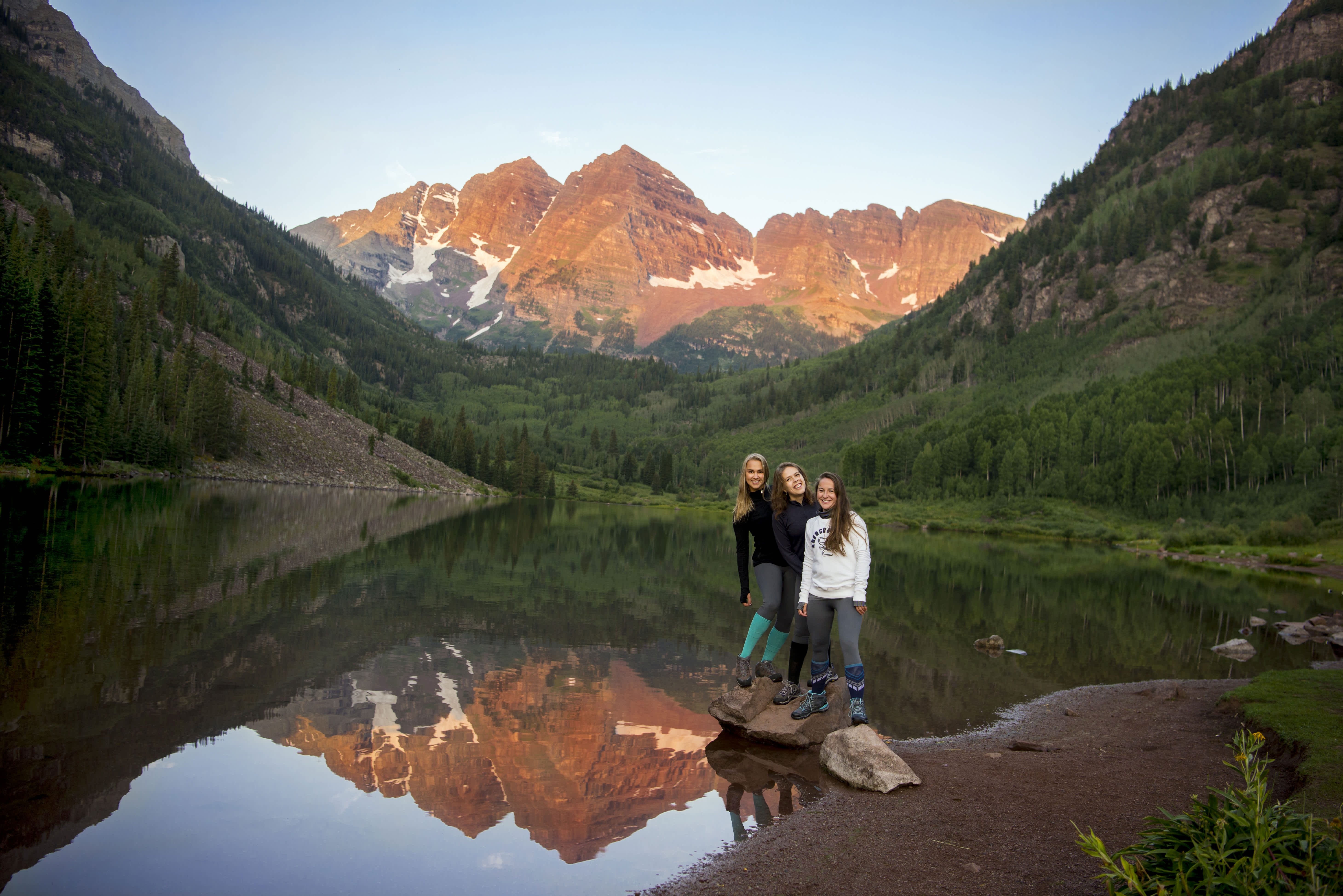 Three women smiling with the Maroon Bells mountain range in the background during sunset.