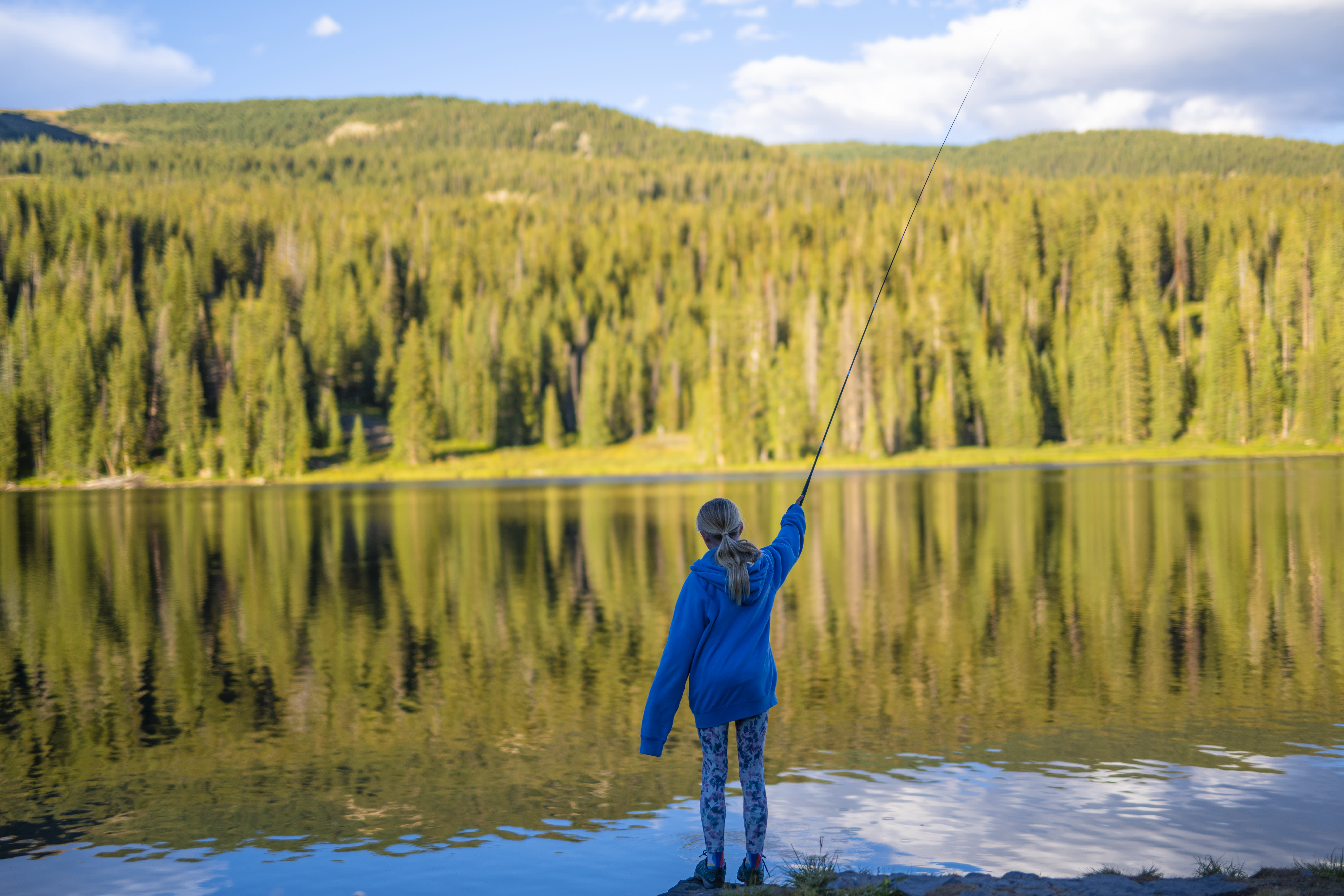 A girl fishing in a forested area in Colorado under a clear blue sky.
