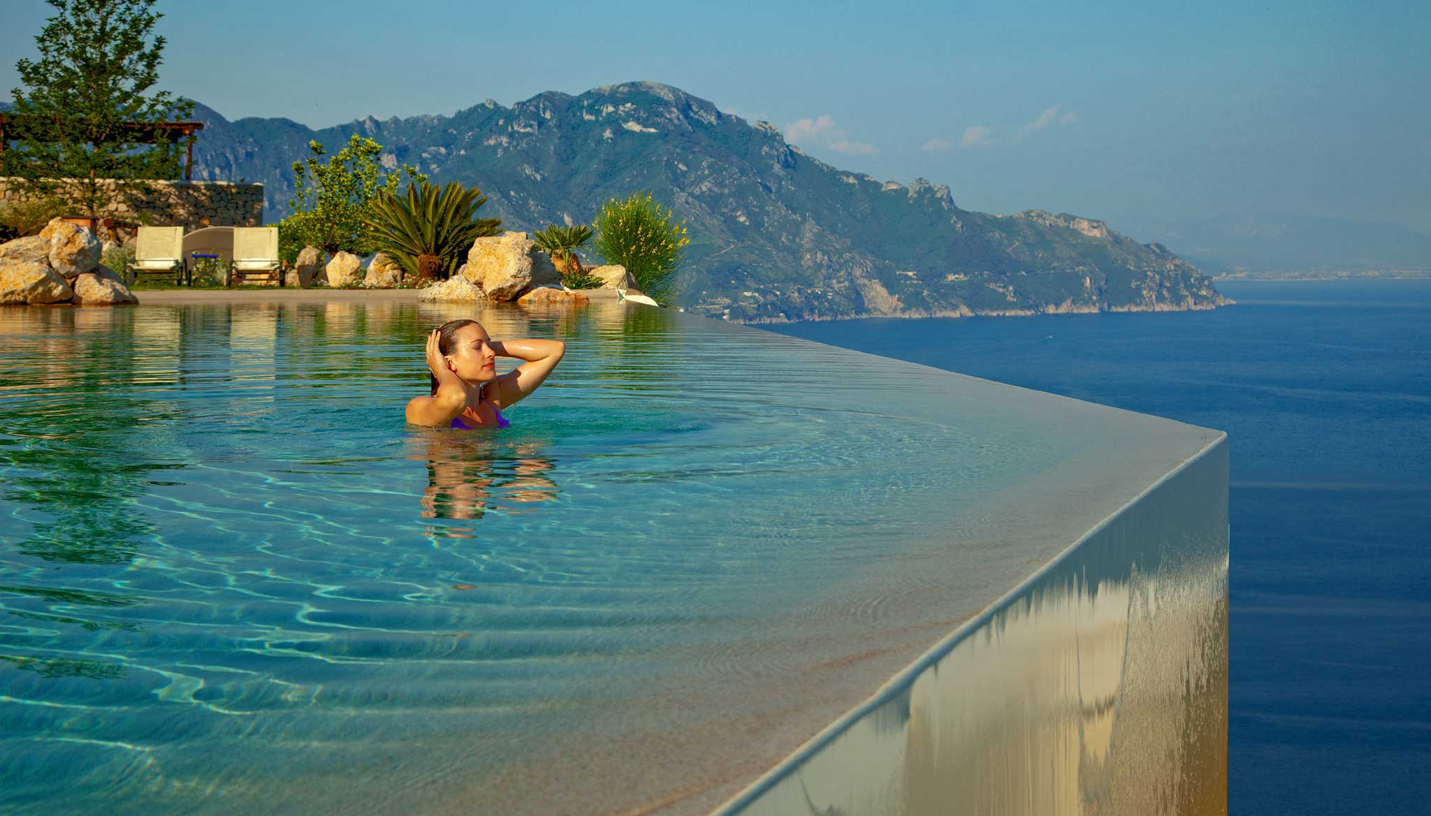 Woman relaxing in an infinity pool with stunning views of the Amalfi Coast and mountains.